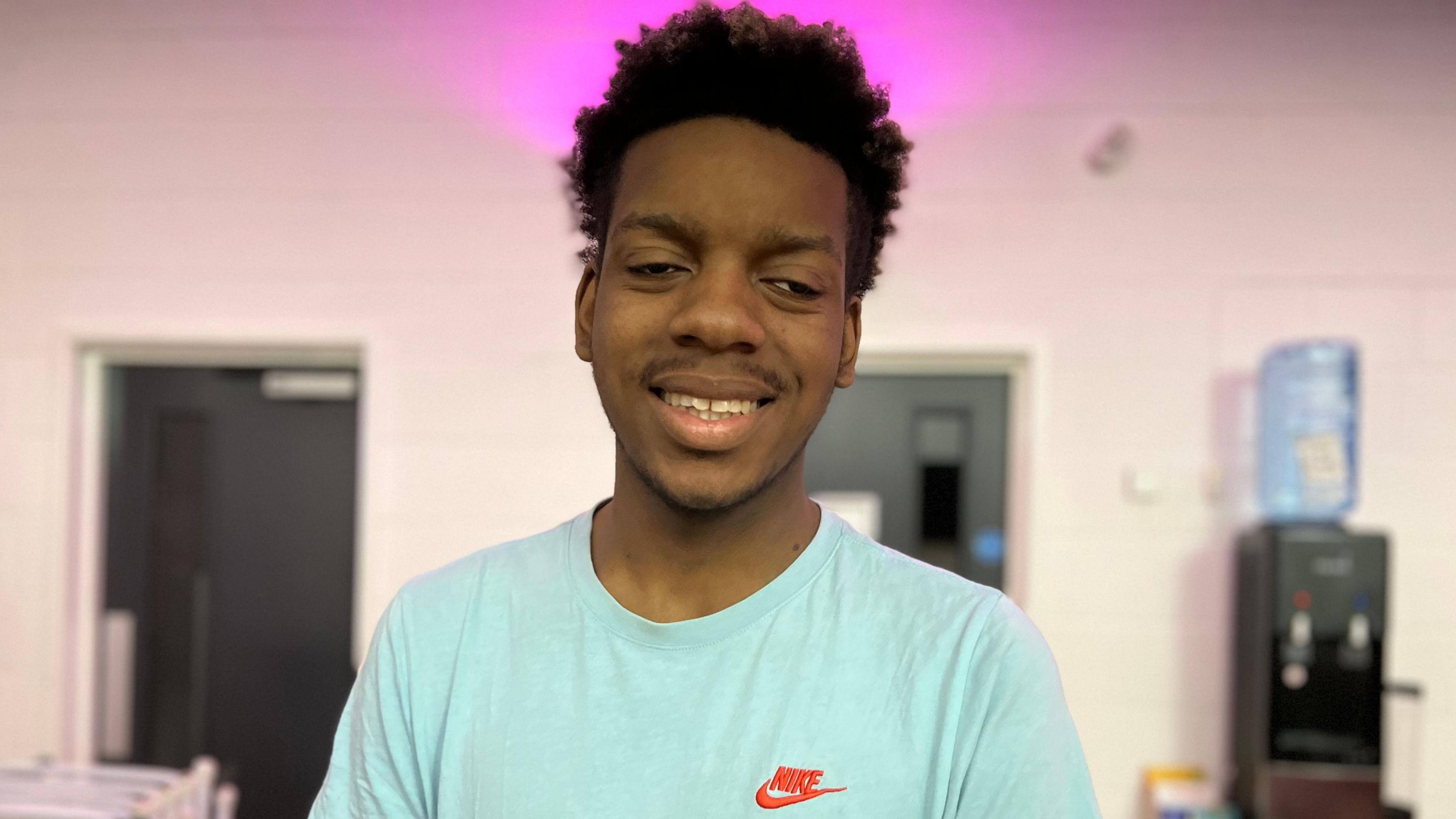A young man with a short afro hairstyle. He is smiling. He wears a light blue T shirt.
