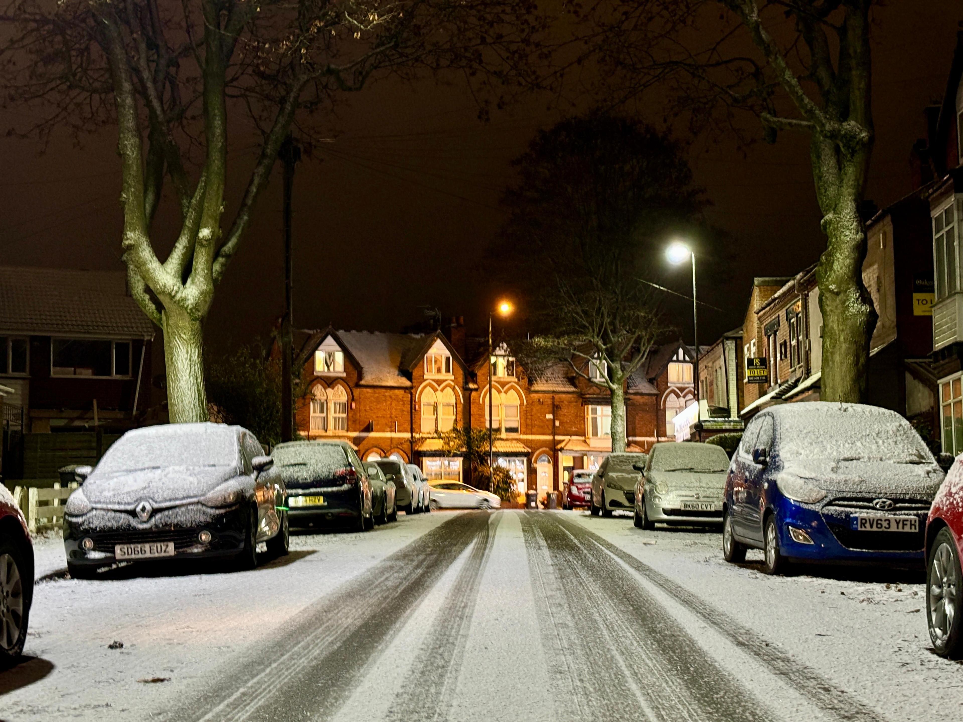 Street-lit buildings are in the centre of this shot but at the rear, with tyre tracks on a snowy road coming towards the photographer and snow-covered cars on either side. 