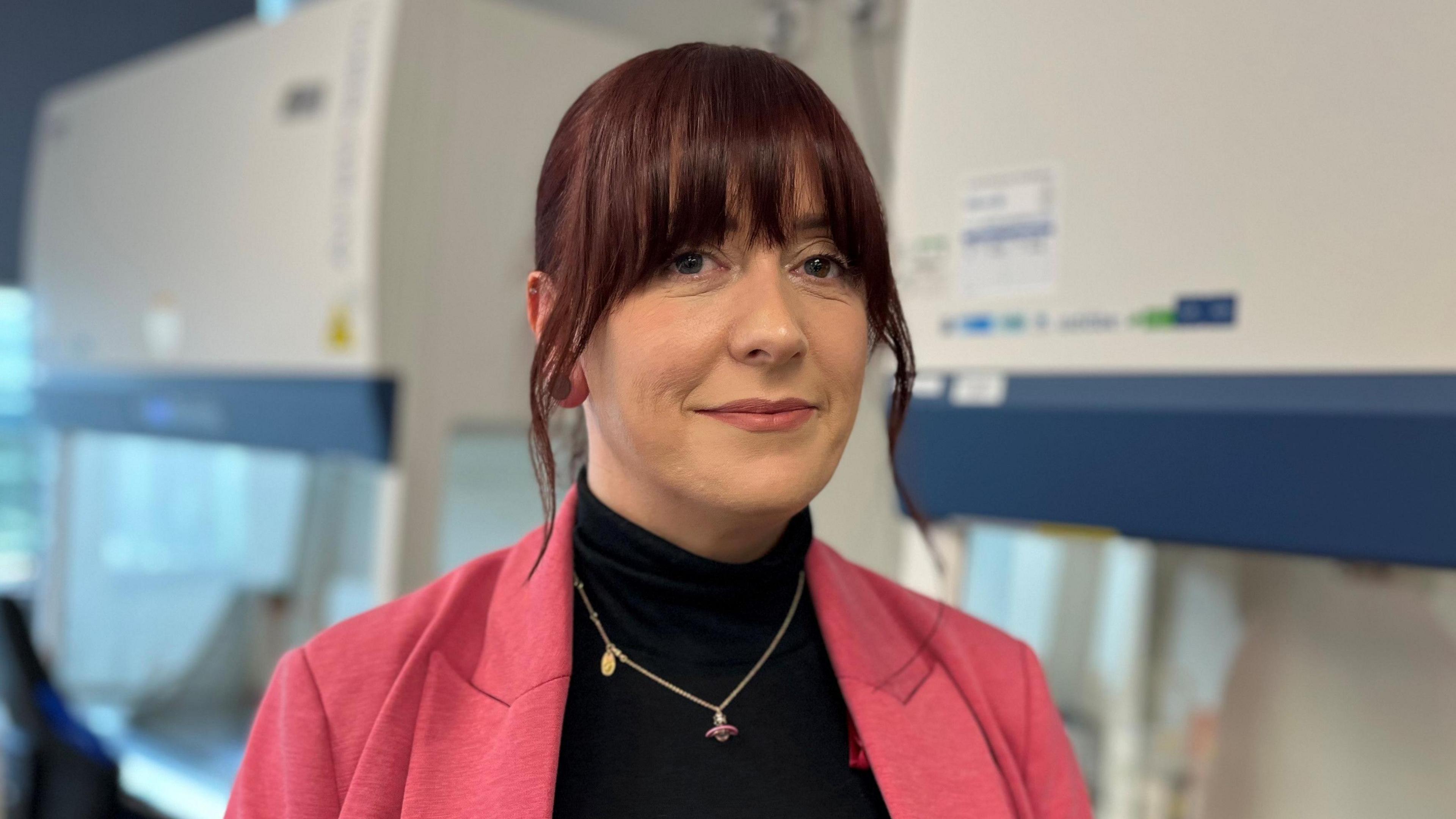 A photo of Lindsay inside a classroom at Falkirk college. She has brown hair with a reddish tint and a strong fringe. She wears a pink jacket and a black jumper with a necklace