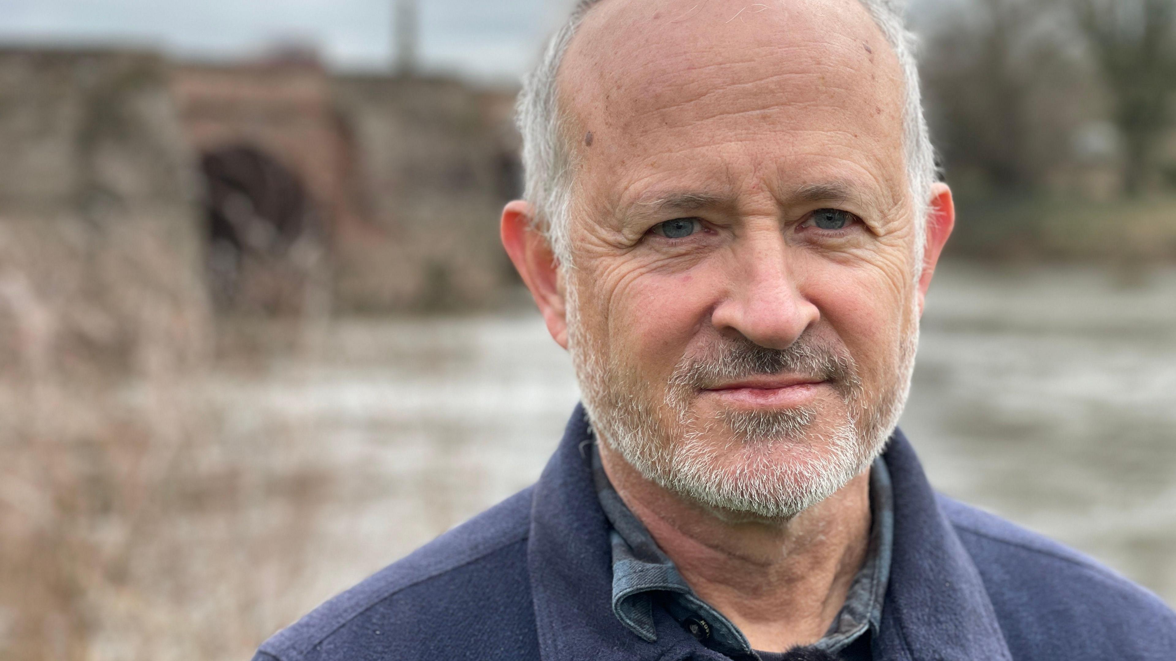 Charles Watson, founder of the charity River Action, stands by the River Wye at Ross on Wye in Herefordshire.  He is wearing a navy shirt. 