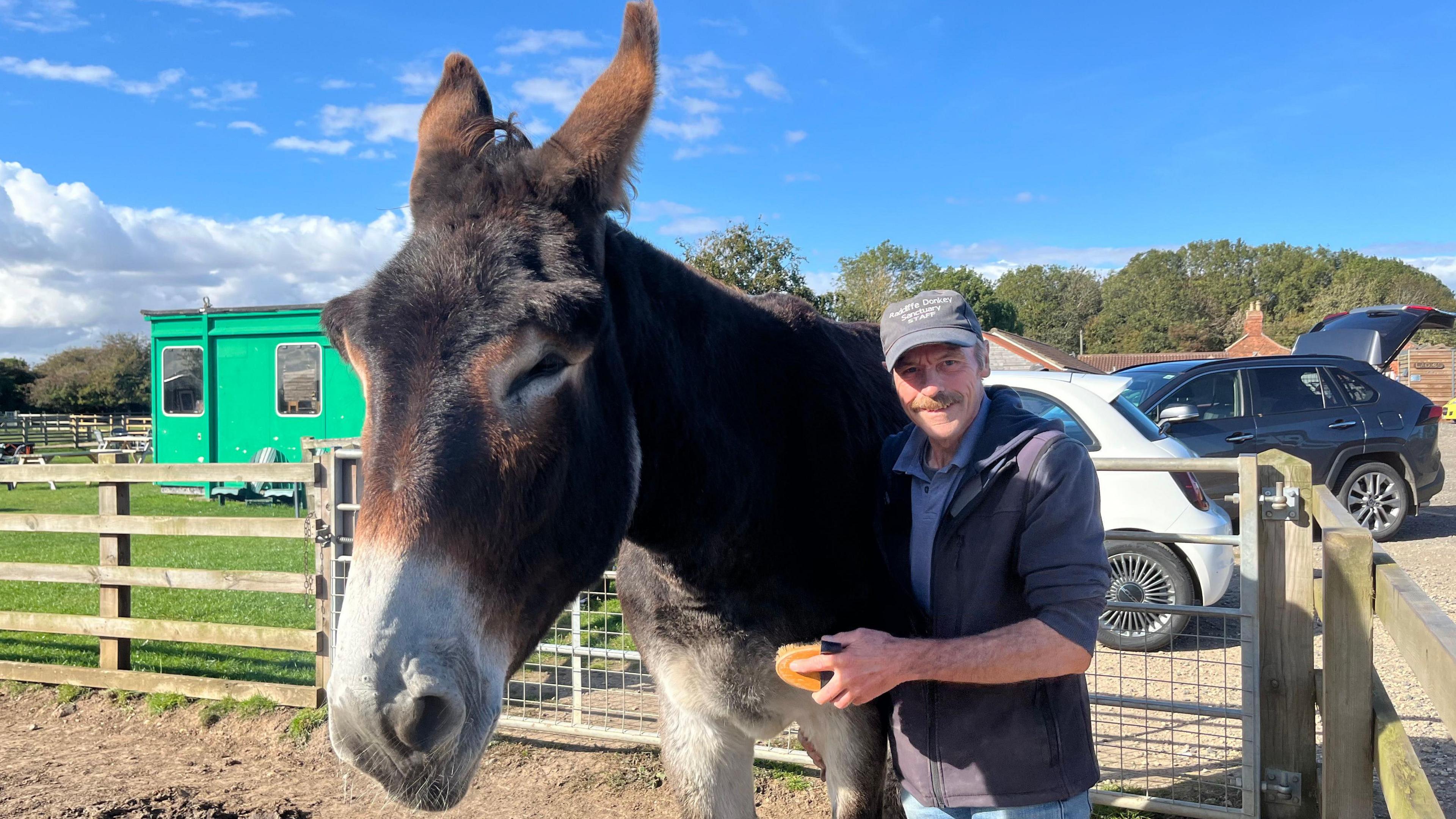 Volunteer Bill Tembey standing next to Derrick. Bill is wearing a cap  and blue jacket and is holding a grooming brush. Derrick's white-tipped nose is in the foreground of the shot. 