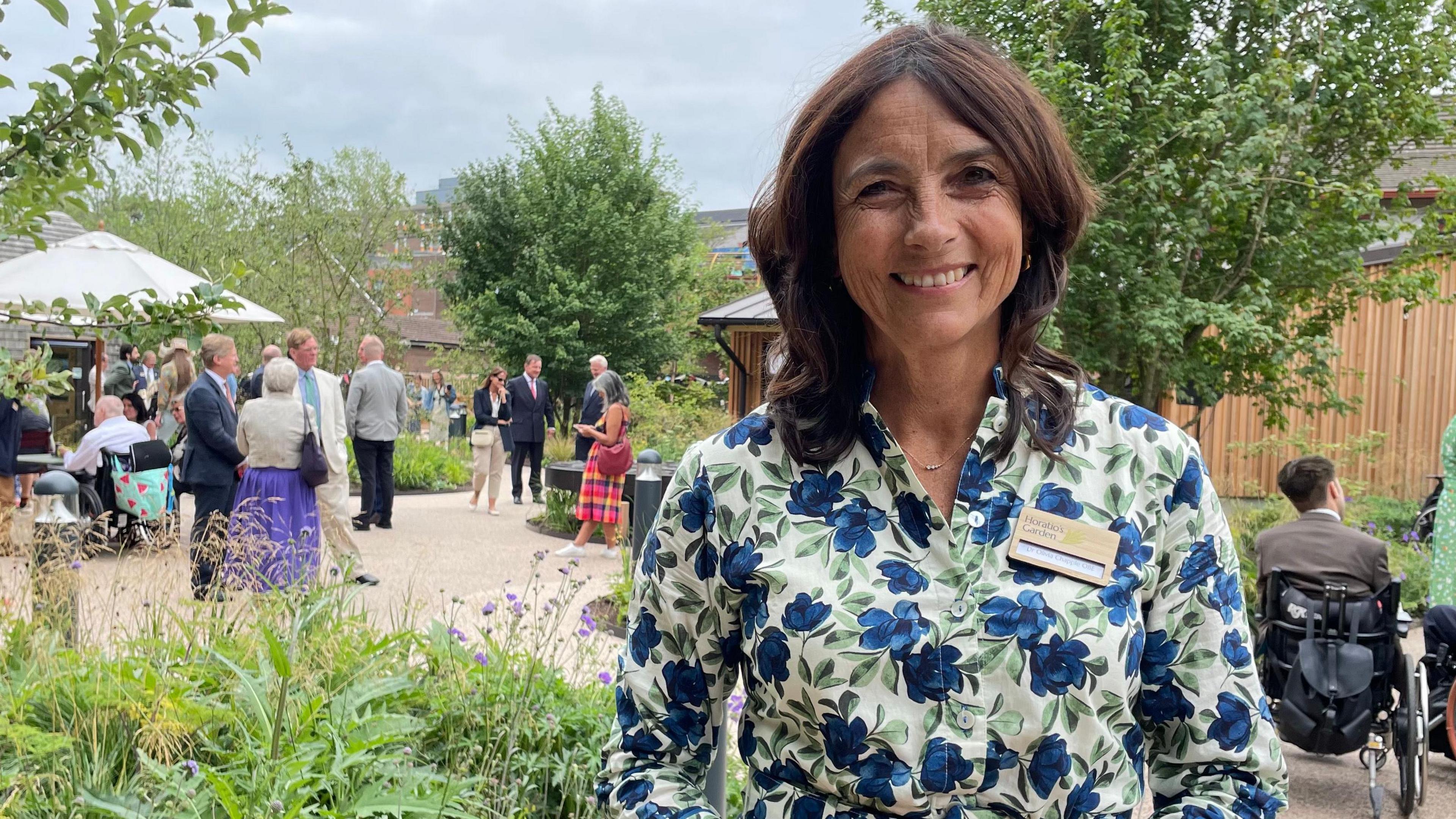 Olivia Chapple, wearing a floral dress, stood smiling in the new garden. Guests of the opening ceremony are stood behind her. One of the defining features of the garden are its wide pathways, which allow for easy wheelchair access.