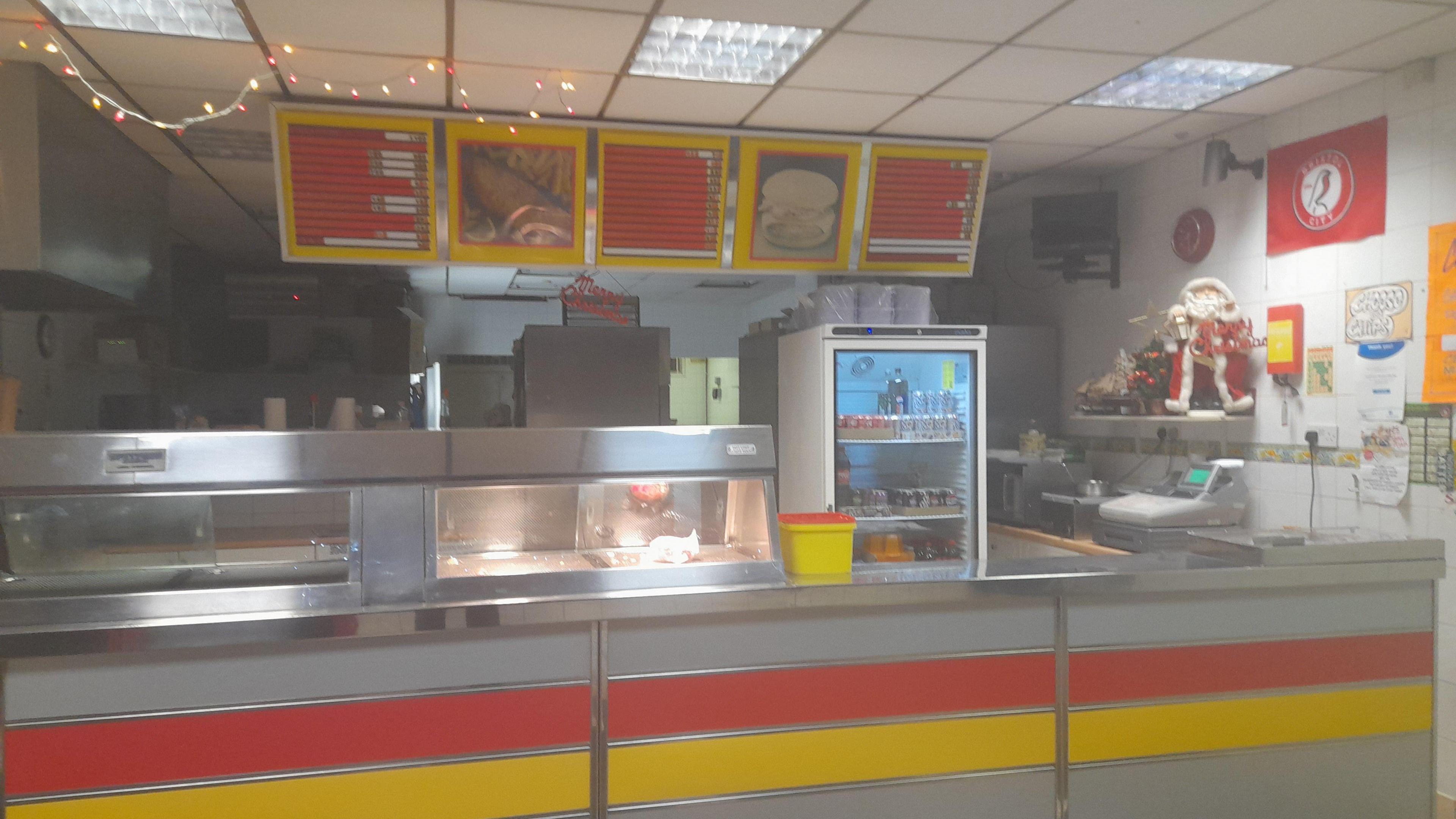 The inside of the chip shop, showing counters and a hot food warmer with a menu board at the top. There is a Bristol City flag on the wall