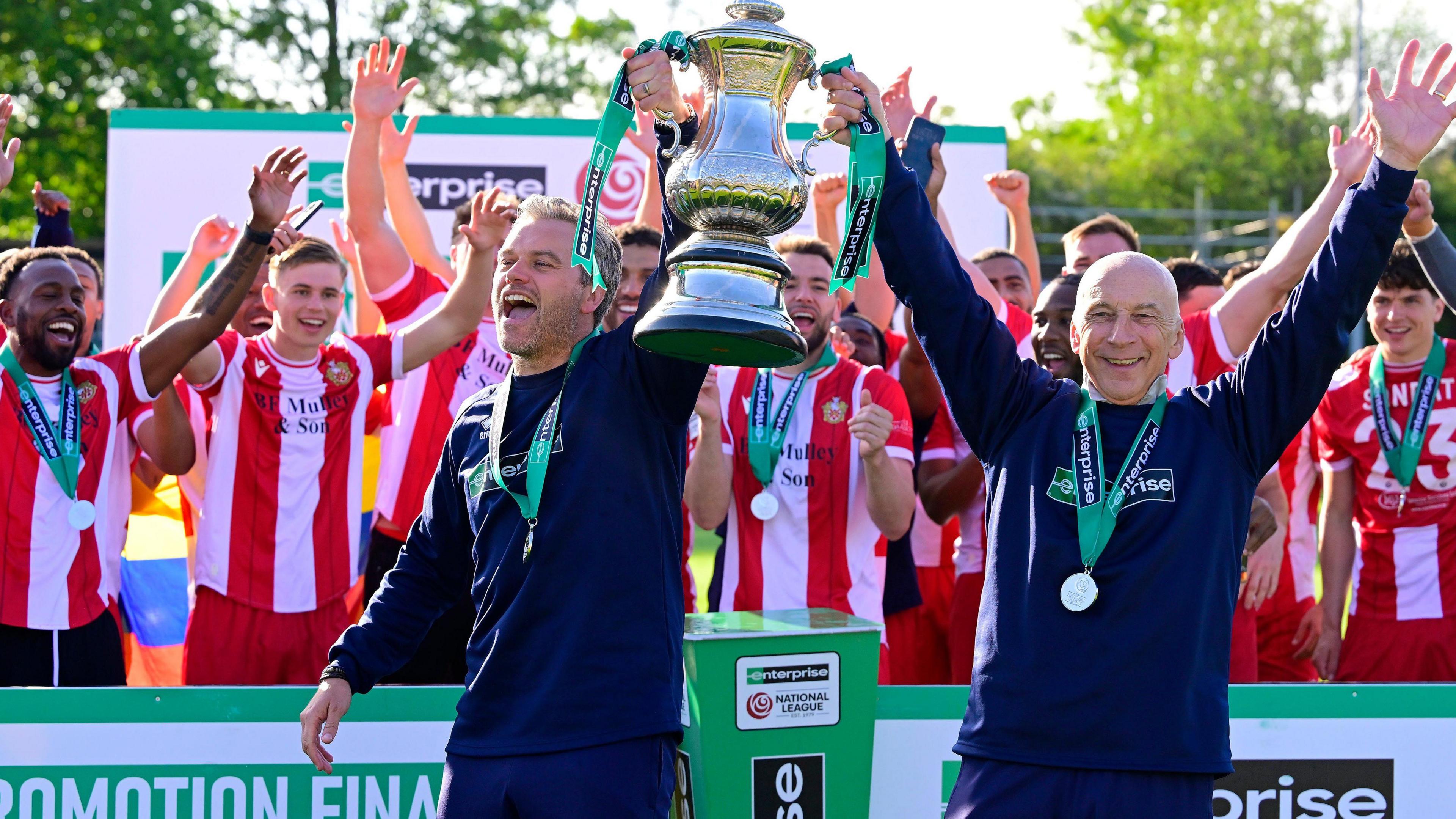 Daryl McMahon, wearing a navy blue tracksuit with a medal around his neck, holds up the National League South play-off trophy with assistant manager Steve Gritt. The Hornchurch players, wearing their red and white striped home kit, can be seen celebrating behind them.