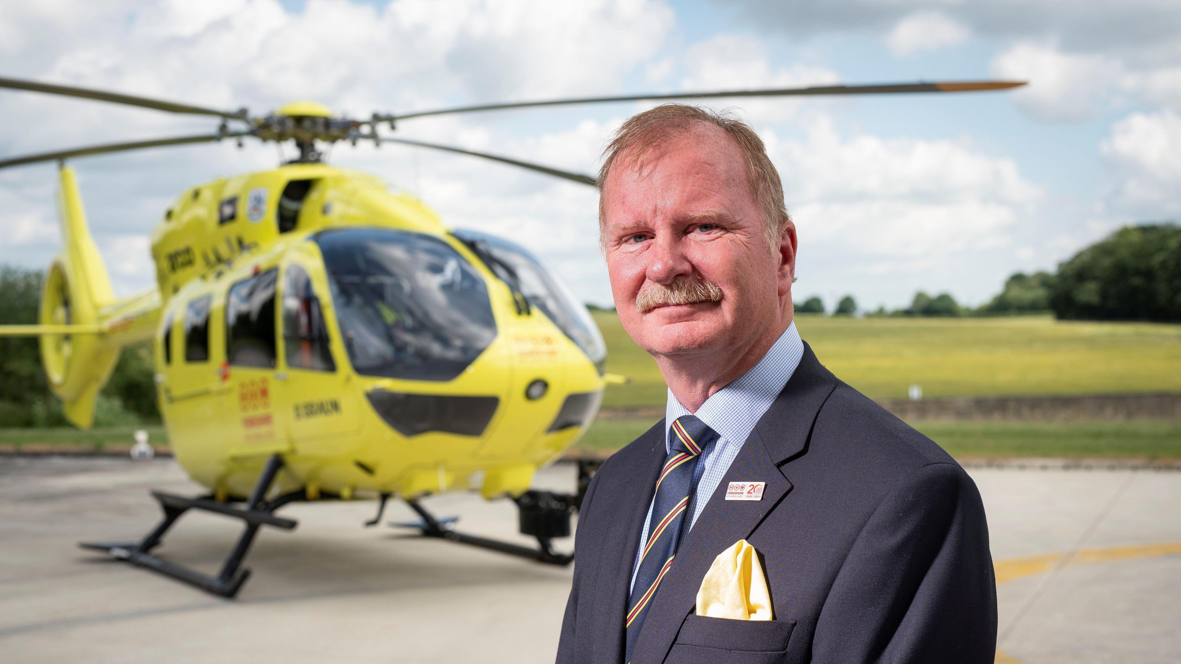 Mike Harrop, chairman of Yorkshire Air Ambulance, with grey hair and moustache is wearing a grey suit and blue striped tie in front of a yellow air ambulance helicopter