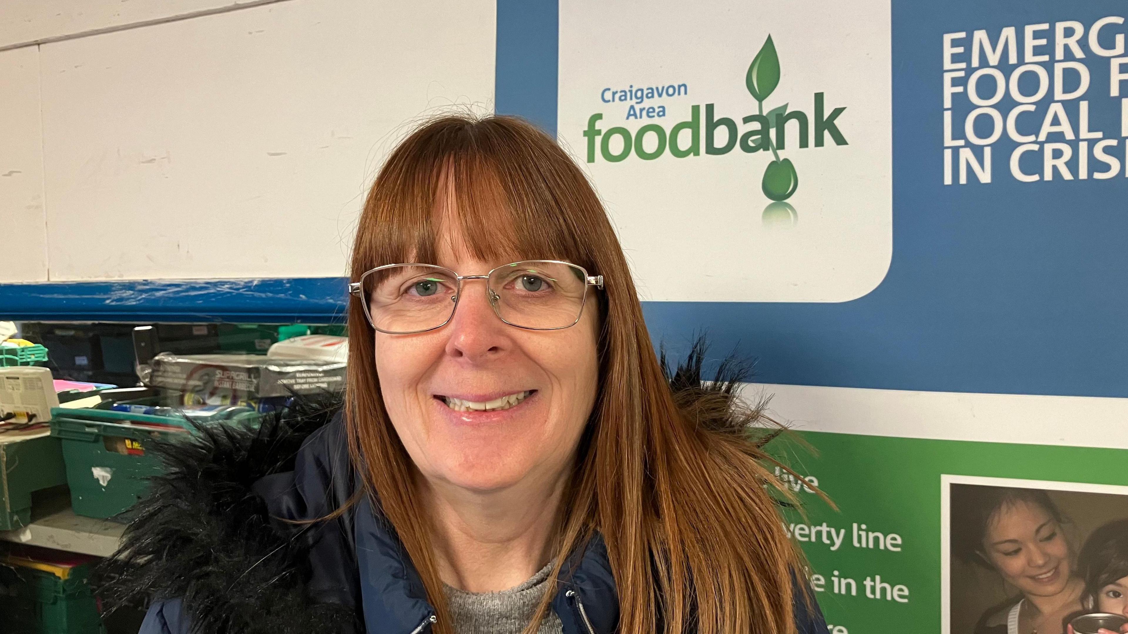 A woman with long auburn hair and wearing glasses is smiling into the camera.  She is wearing a dark navy coat with fur around the hood. Behind her is a green light blue and white sign for Craigavon Area foodbank.

