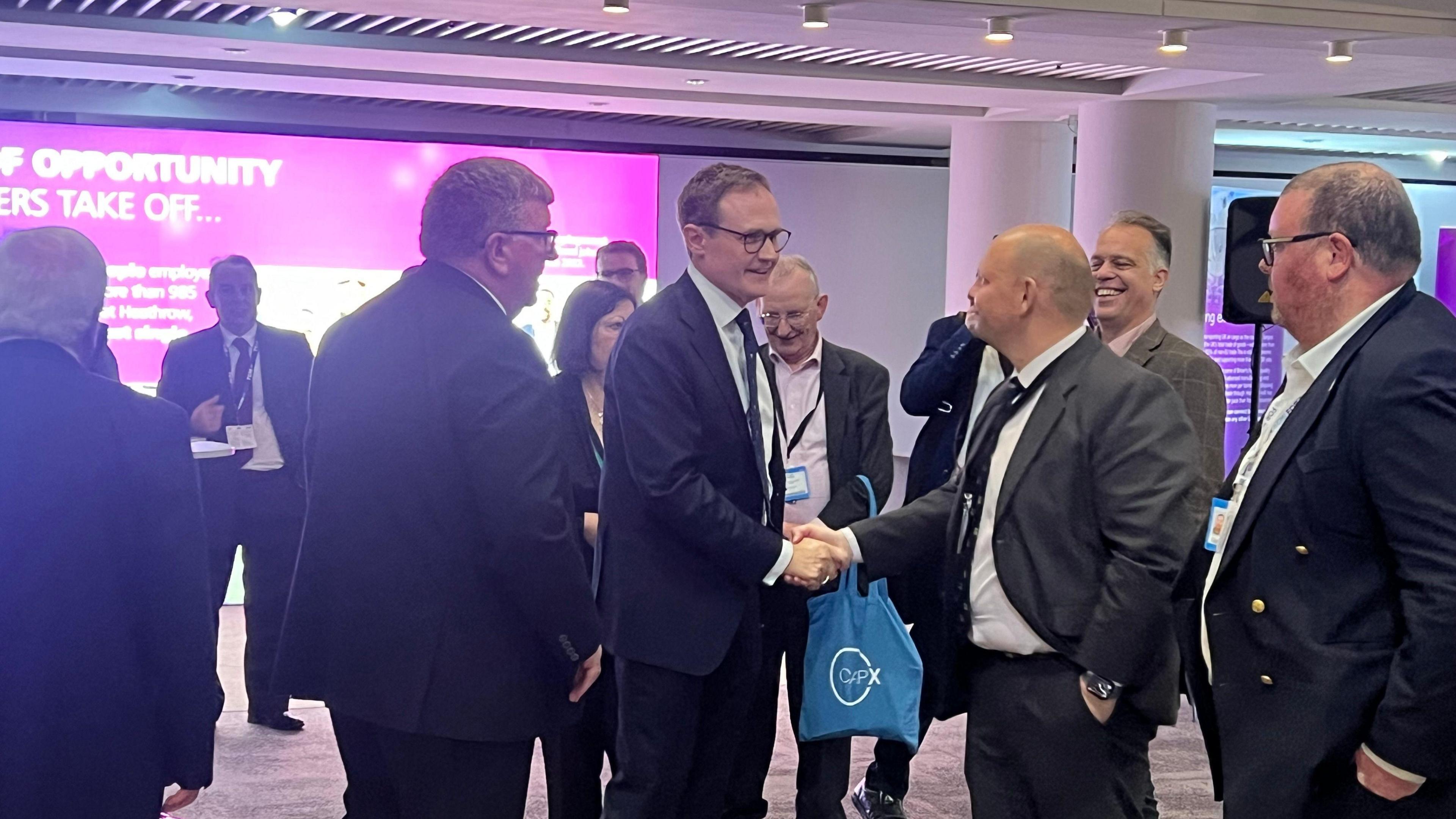 A man with glasses and a suit smiles as he shakes hands with another man in a suit at the tory conference. They are surrounded by other men attending the conference. 