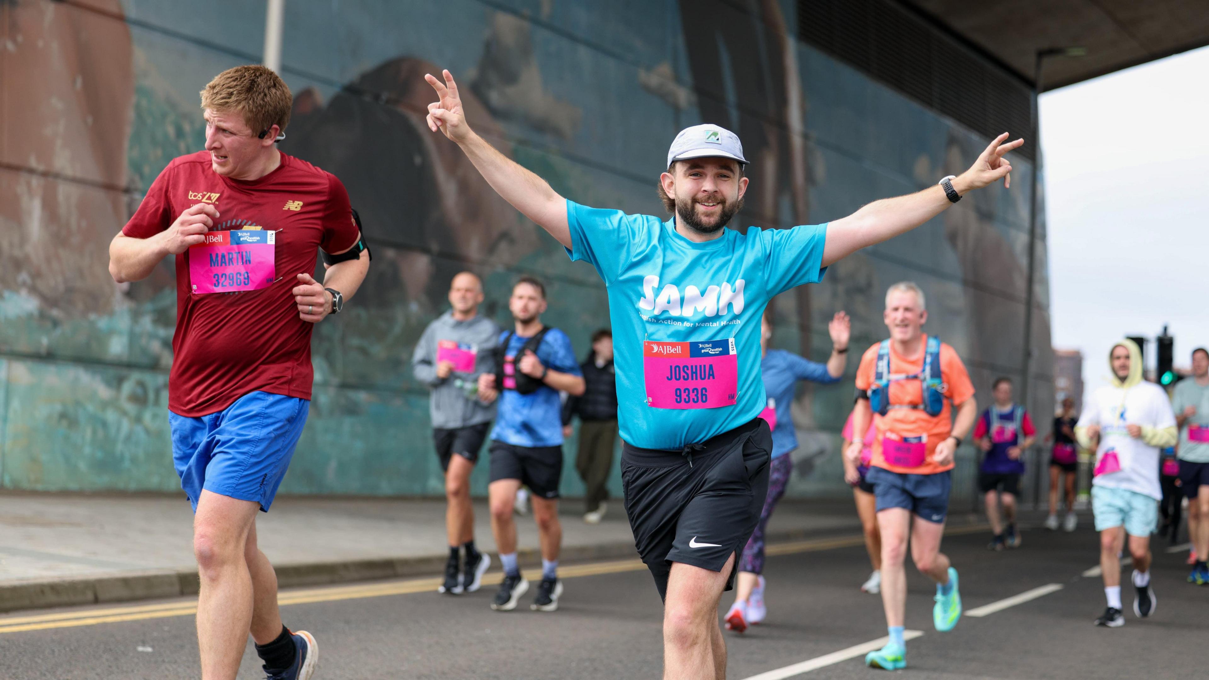 a runner wearing a blue "SAMH" shirt and a white cap, makes peace signs with both hands.