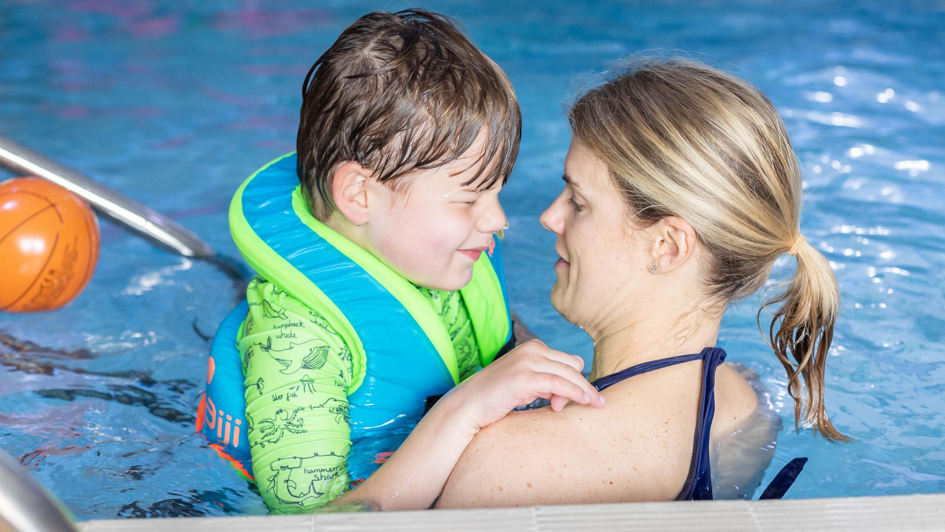 A woman in a swimming pool looking at a chid, who she is carrying. The child is wearing bright green and blue swimming gear, including a float jacket. An orange ball is floating in the background.