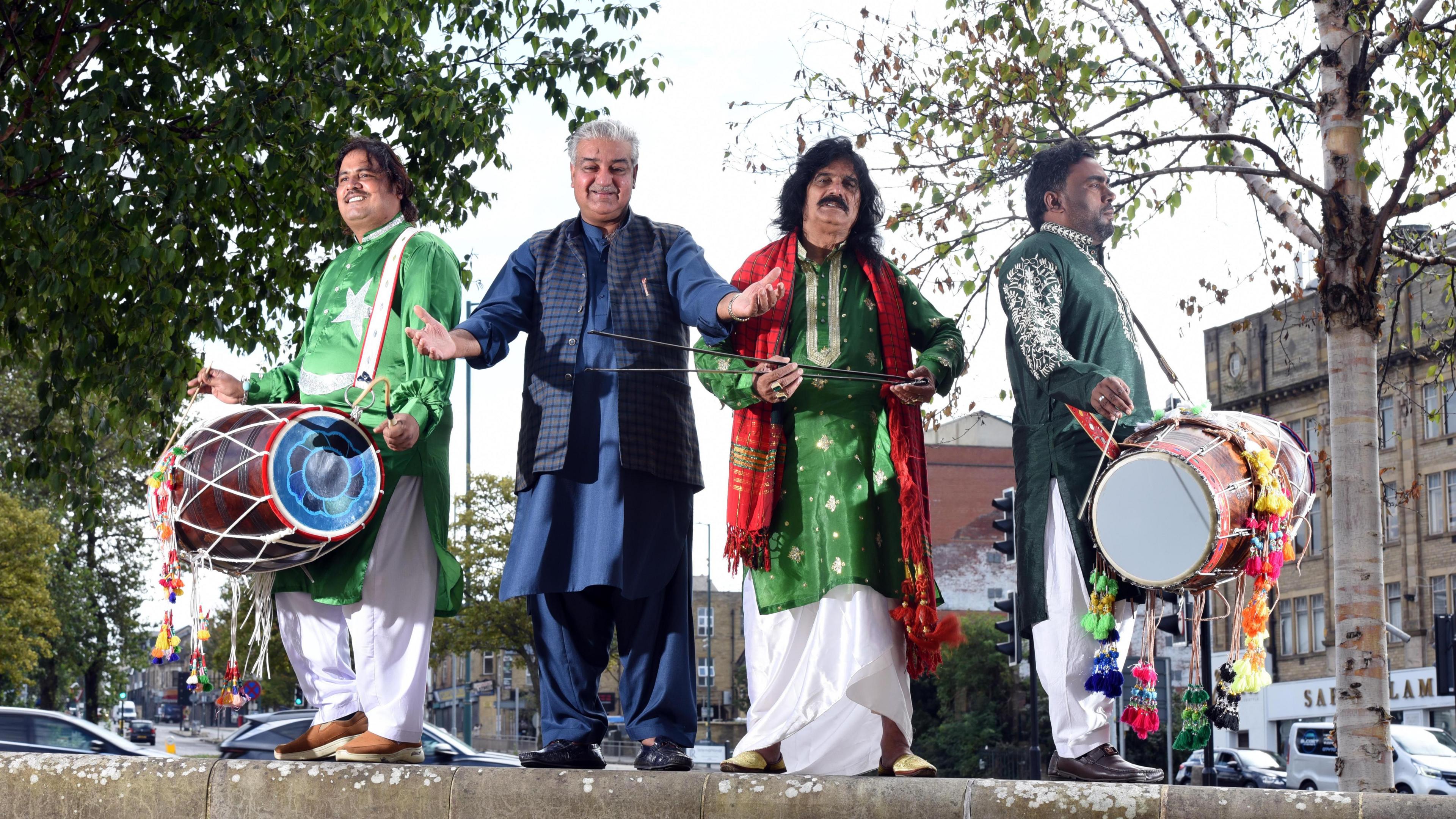 Four South Asian men pictured in an urban streetscape. They are dressed in traditional clothes. Three of them carry dhol drums and other musical instruments.