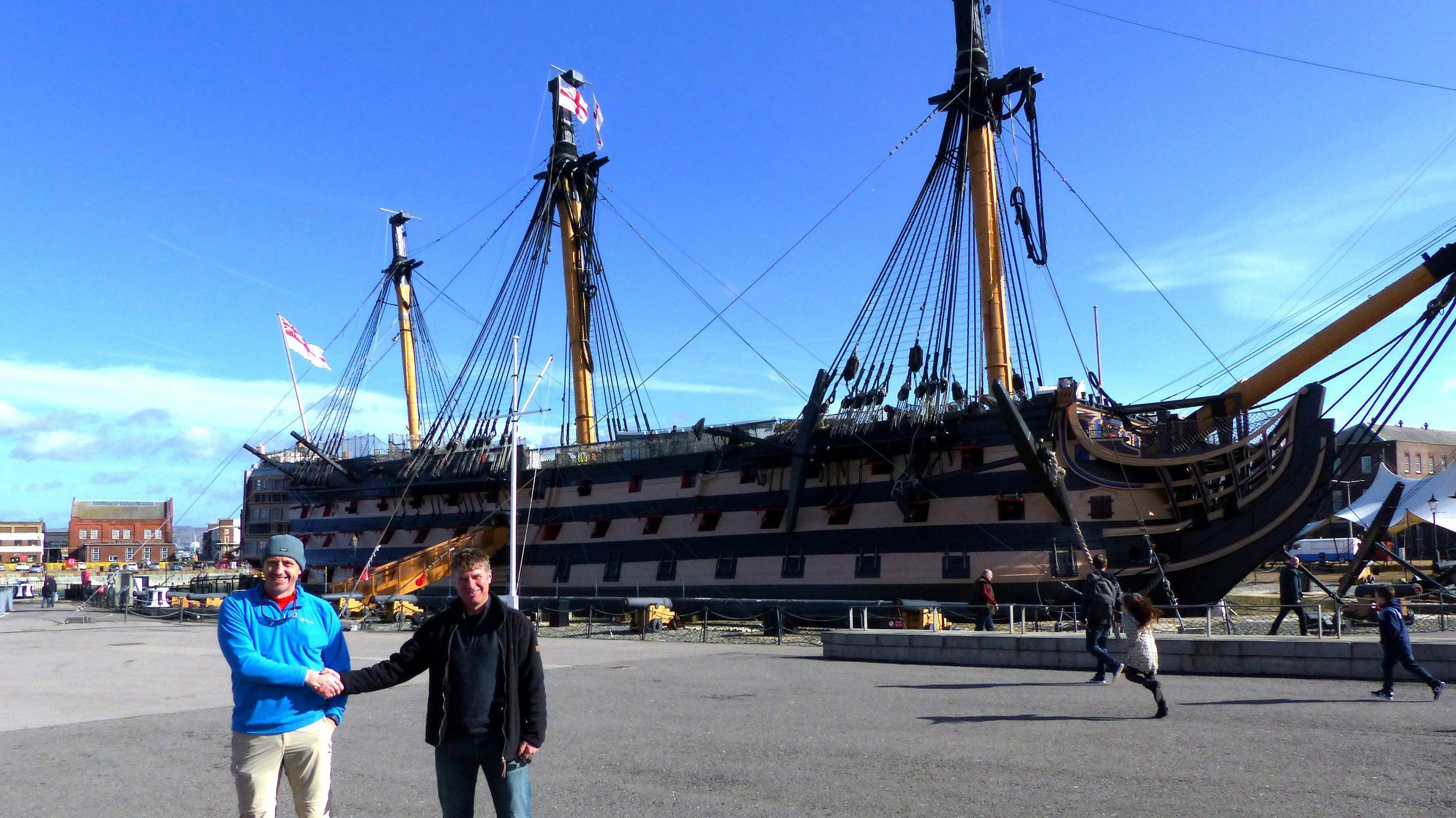 Two men shaking hands. They are standing in front of a large vessel moored at Portsmouth harbour.