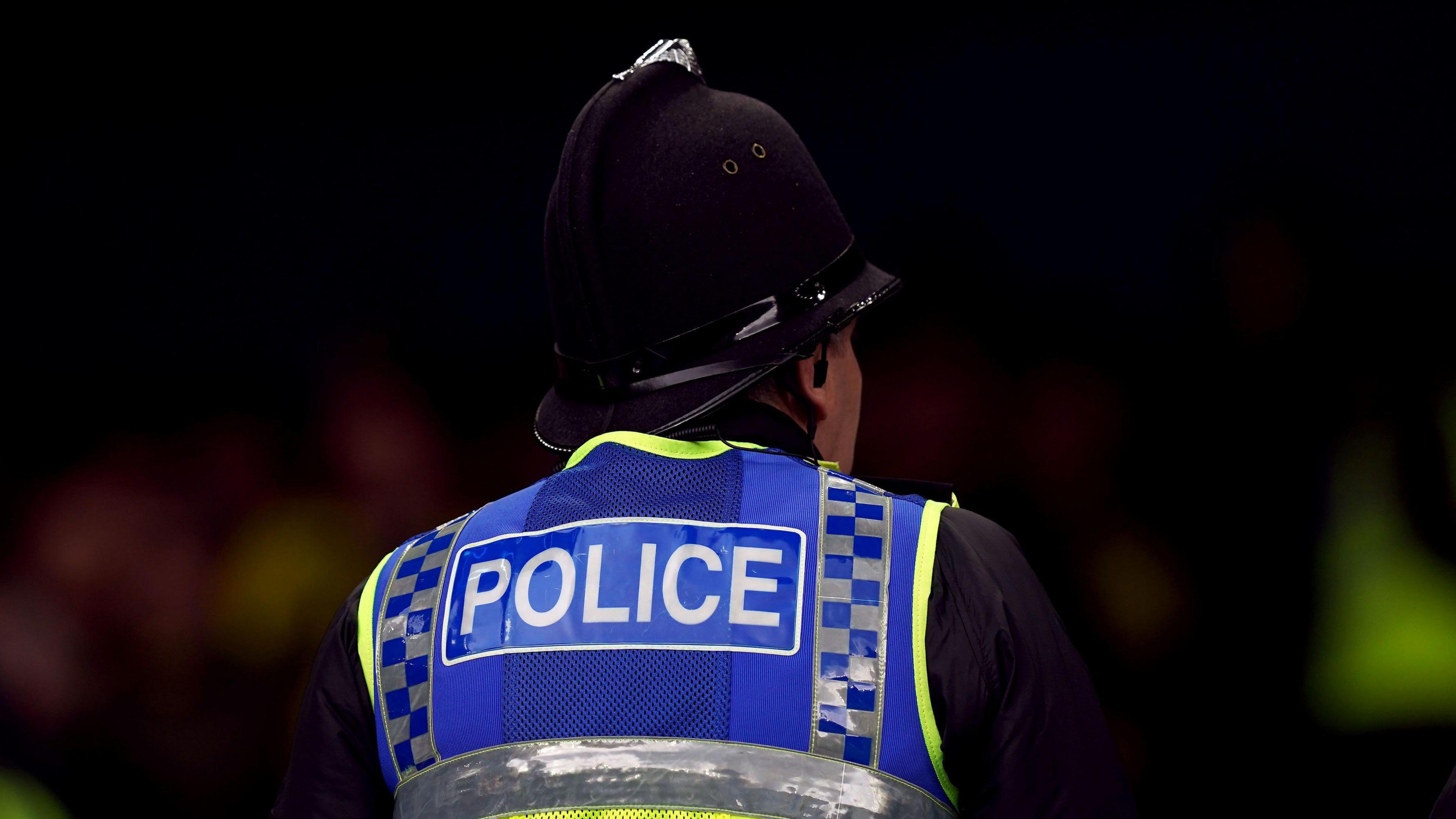 A police officer wearing a hi-vis vest and a traditional hat with his back to the camera