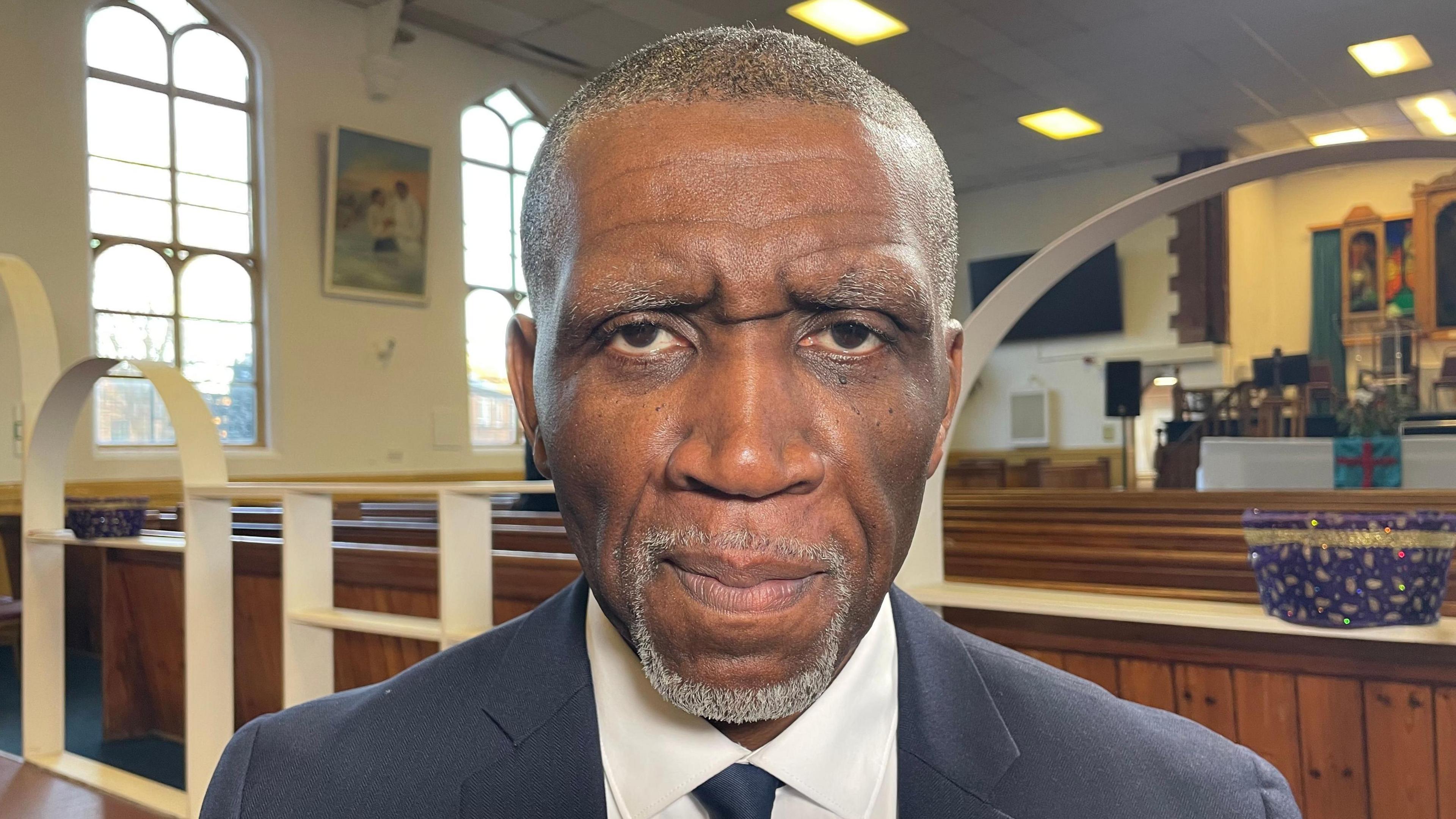 Clive Foster is wearing a suit and tie and standing in front of church pews