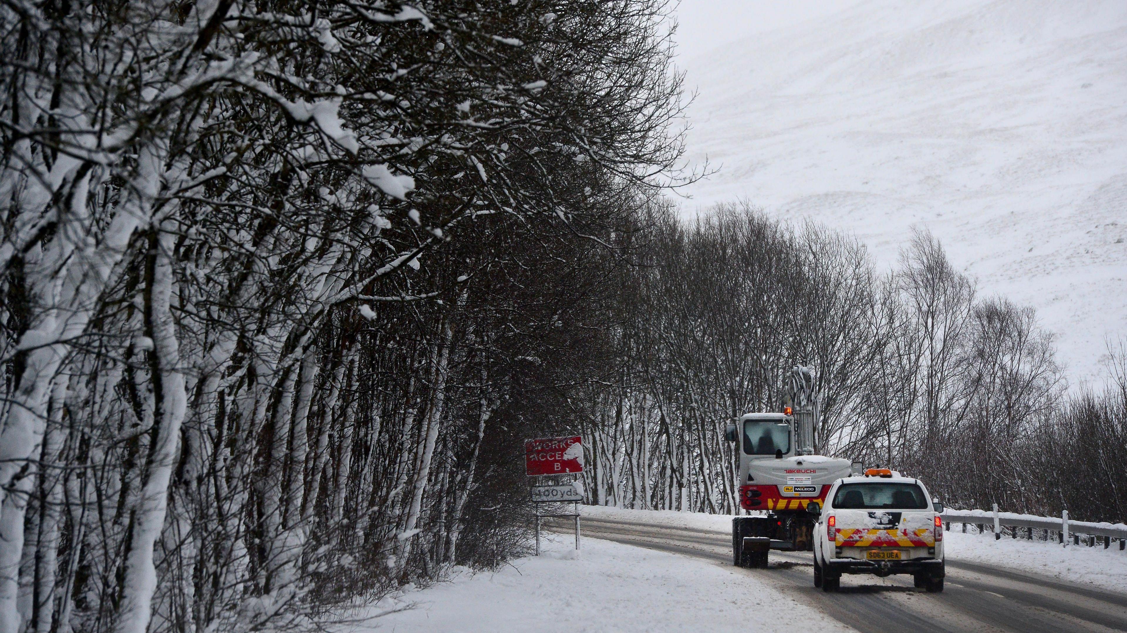 A covering of snow and ice on a hillside road with a snow plough followed by a safety vehicle. Snow-covered hill and grey sky in the background.