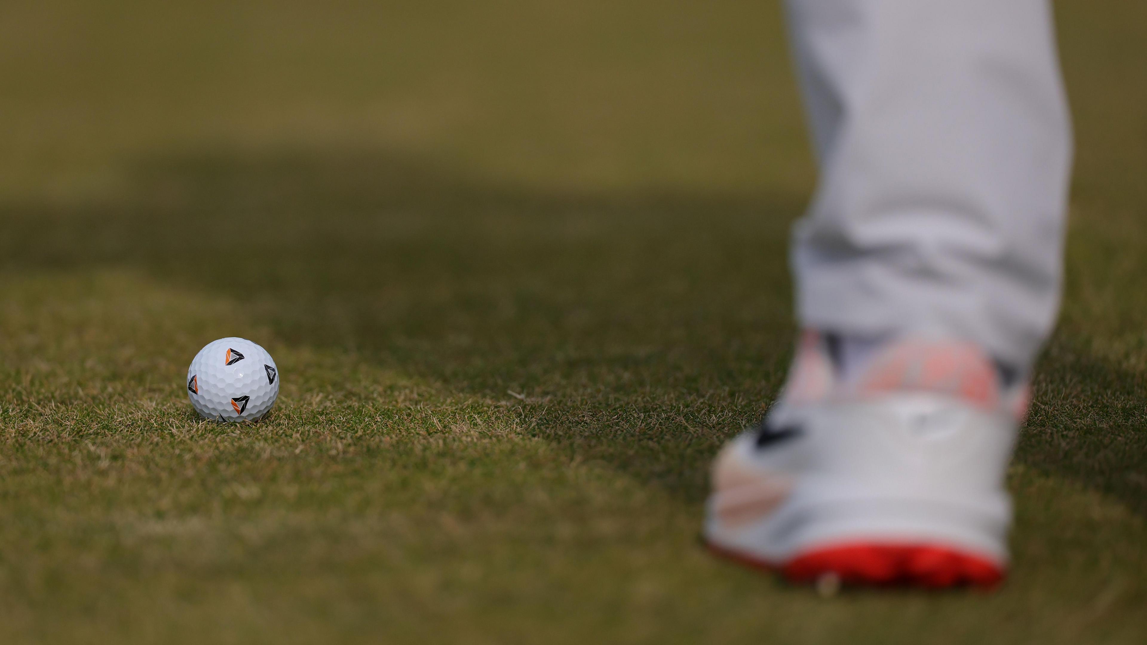 Close up of a white golf ball with four distinctive triangle pen markings. A blurred white golf shoe, with orange soles, is blurred in the foreground