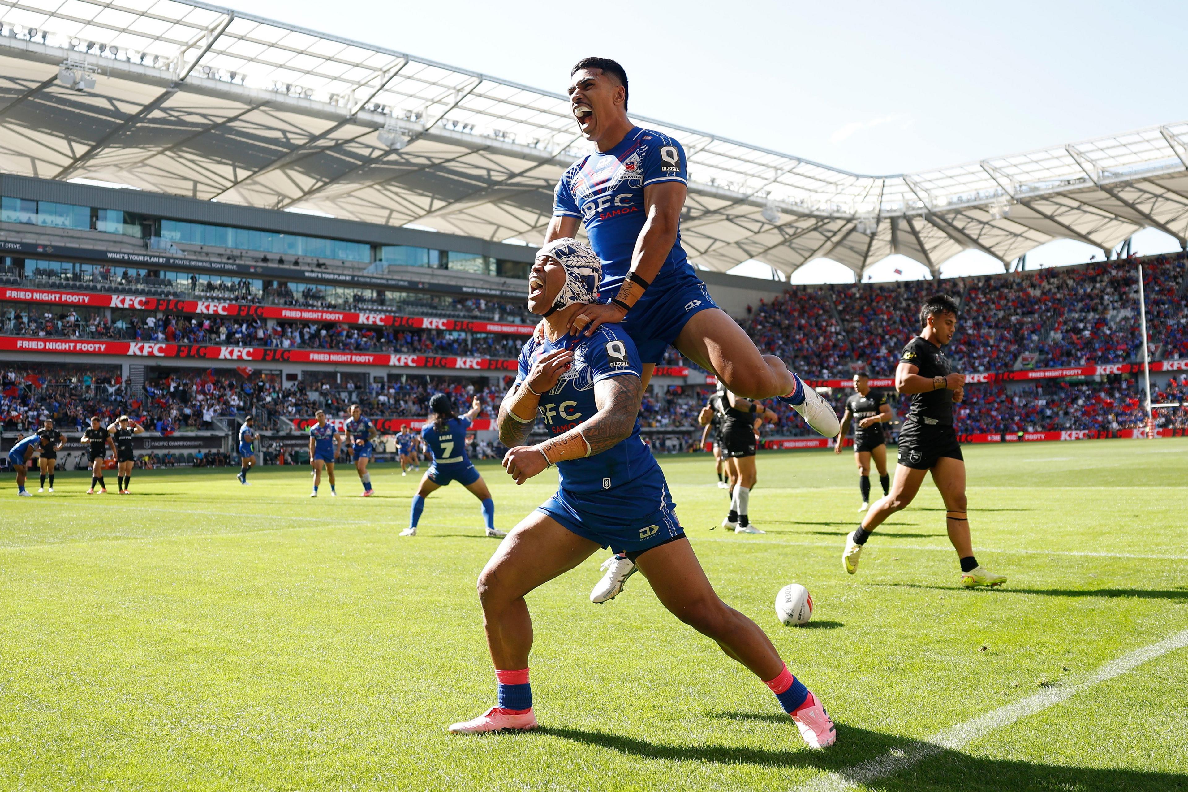 Two rugby players in blue jerseys celebrate on the field; one player is lifting the other onto their shoulders. The scene takes place in a stadium with a large crowd in the background and other players visible on the pitch.