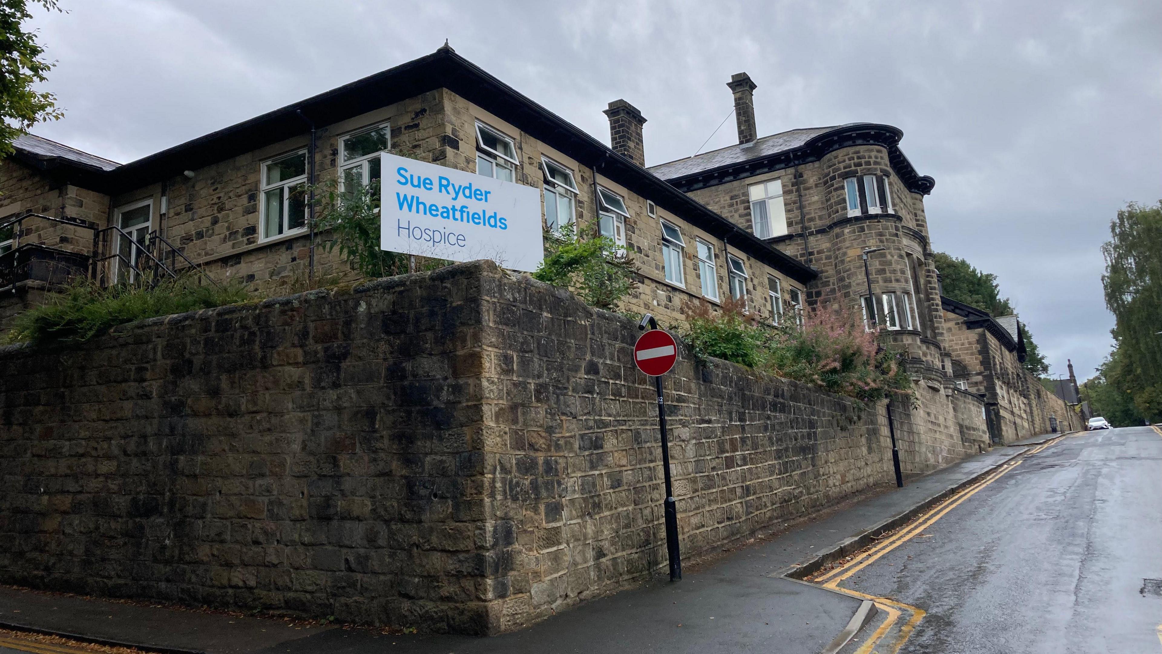 A stone building on the corner of a road with a sign outside which reads 'Sue Ryder Wheatfields Hospice'.