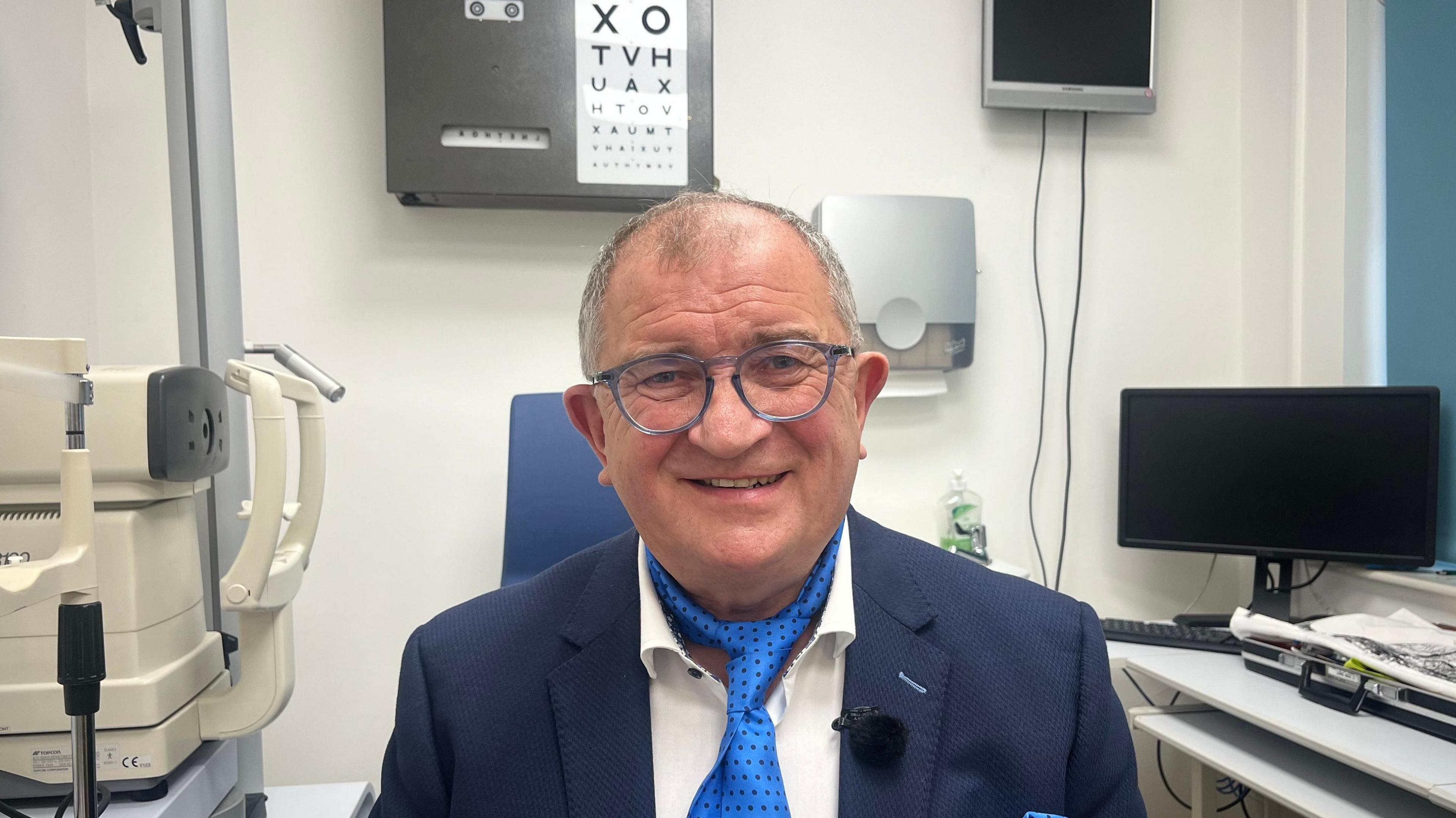 A man wearing a blue suit and blue neck tie is smiling at the camera. Behind him is some medical equipment.