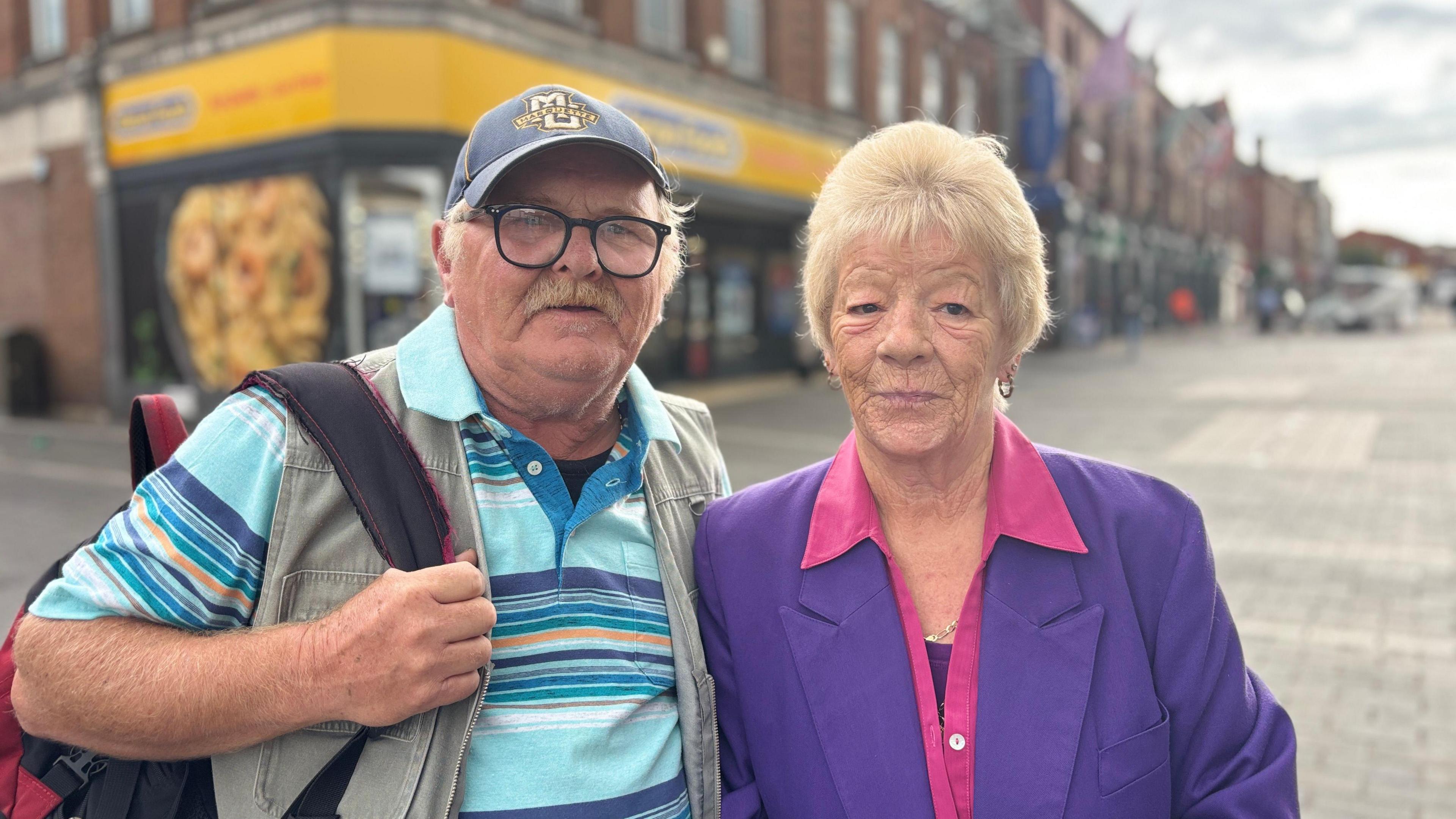 A man wearing a blue striped t shirt and a grey waistcoat is on the left of the picture. He is also wearing glasses and a baseball cap and carrying a rucksack. 
A lady wearing a purple coat and pink blouse is stood on his right hand side. 
Behind them is a street scene including Heron foods with a yellow sign. 