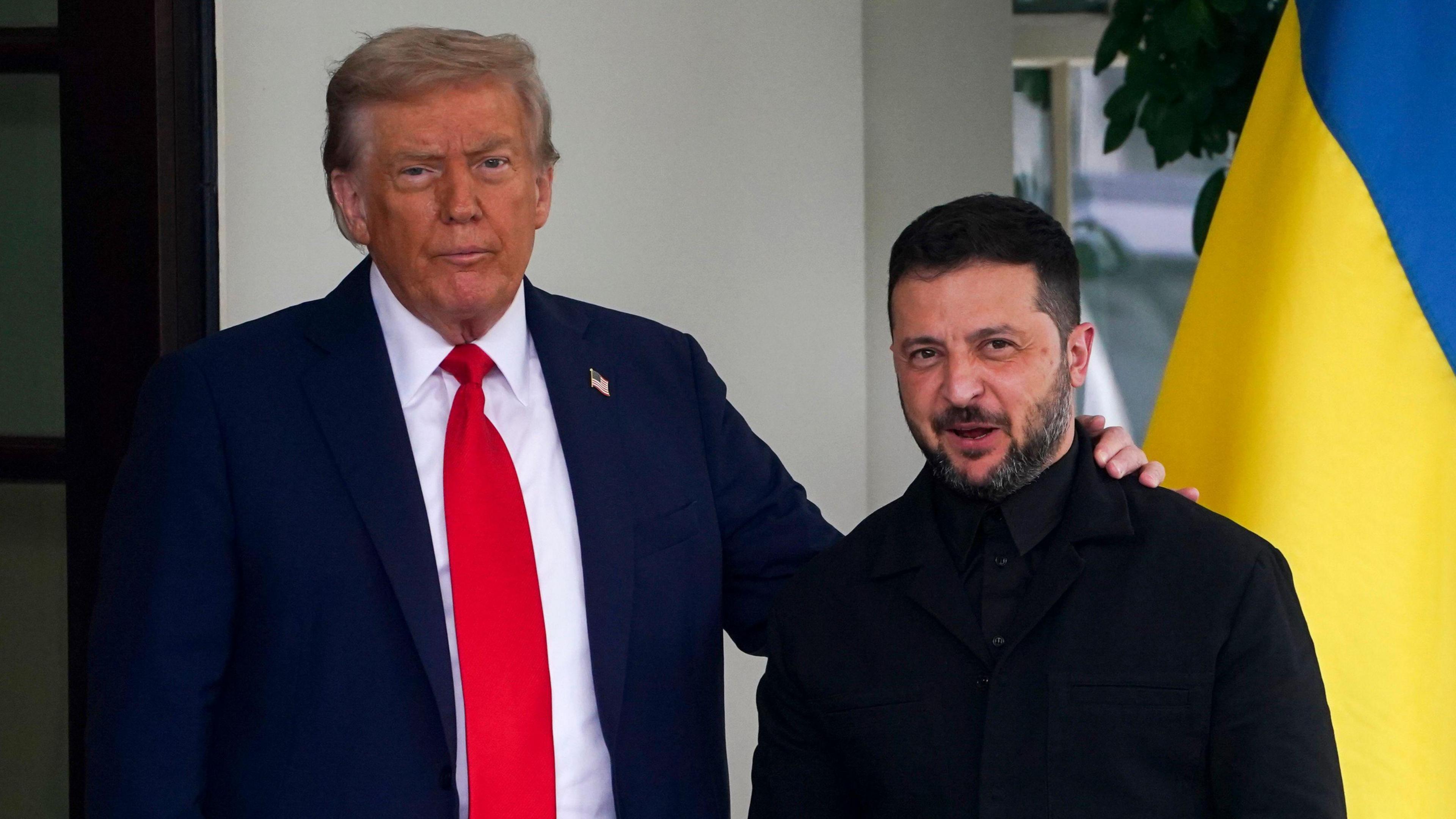 Donald Trump, wearing a blue suit and red tie, places a hand on the shoulder of Volodymyr Zelensky, dressed in black, with a Ukrainian flag visible behind them