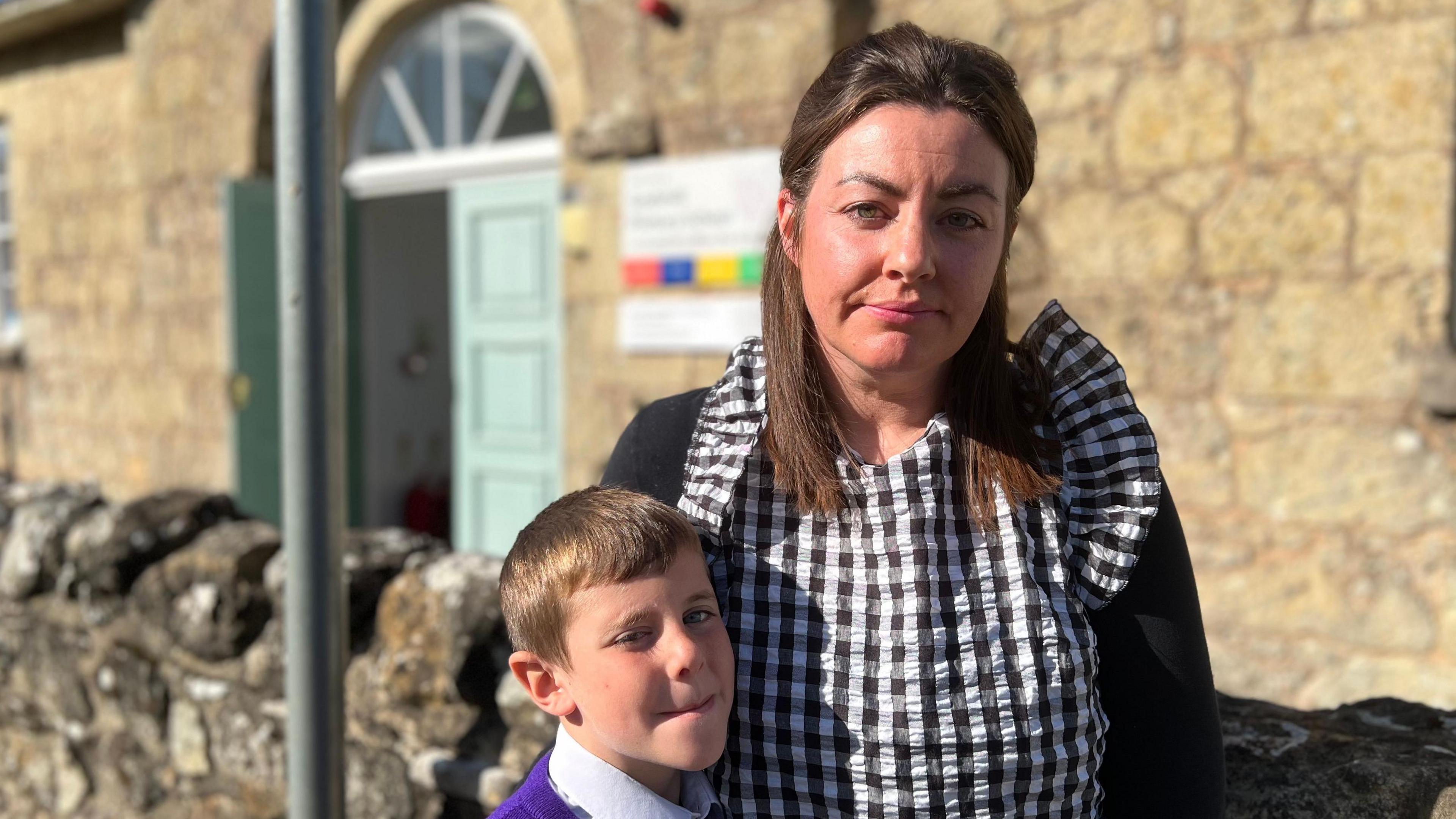 A mum wearing a black and white checked stop with her son in purple school uniform stood outside Godshill primary school. They are both looking sad.