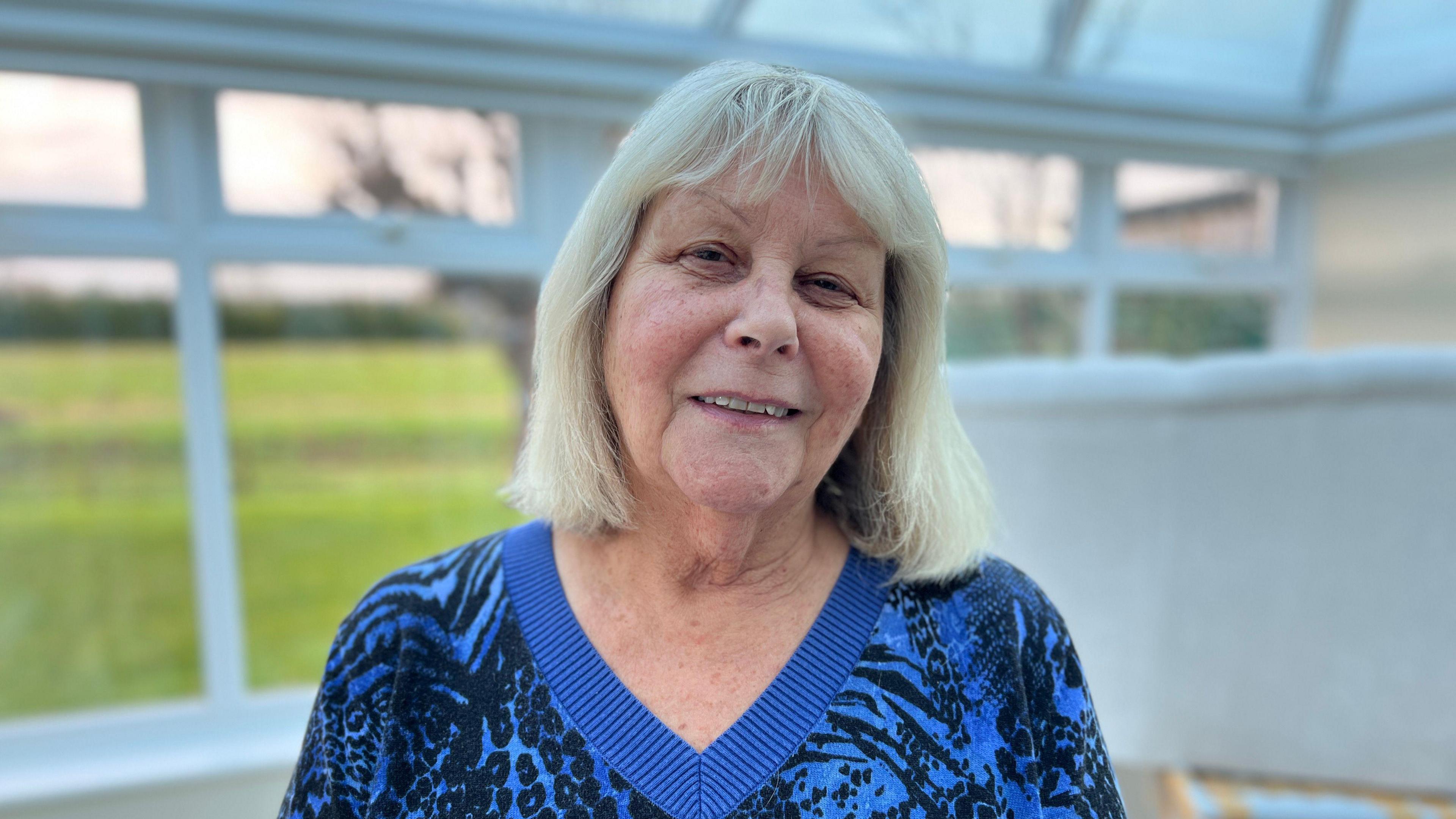 Dauntsey resident Jacque smiles at the camera. She has white hair to her shoulders and is wearing a blue top with a v-shaped neck. She is standing in a conservatory. 