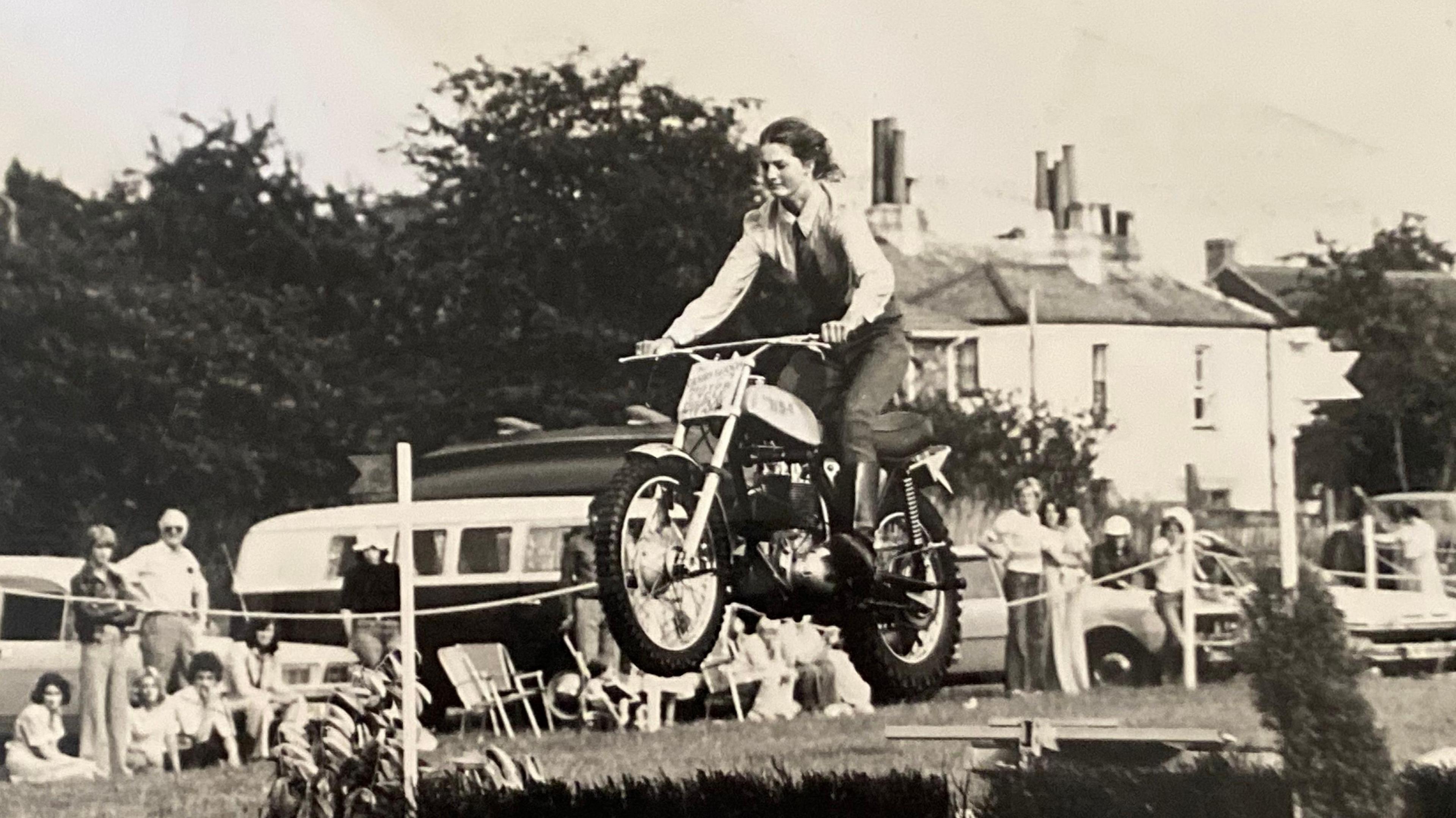 A black and white archive photo showing a woman riding a motorcycle over an equestrian-style jump at a village fete-style event. In the background, people watch from behind a cordon, which is stretched in front of parked cars and camper vans.