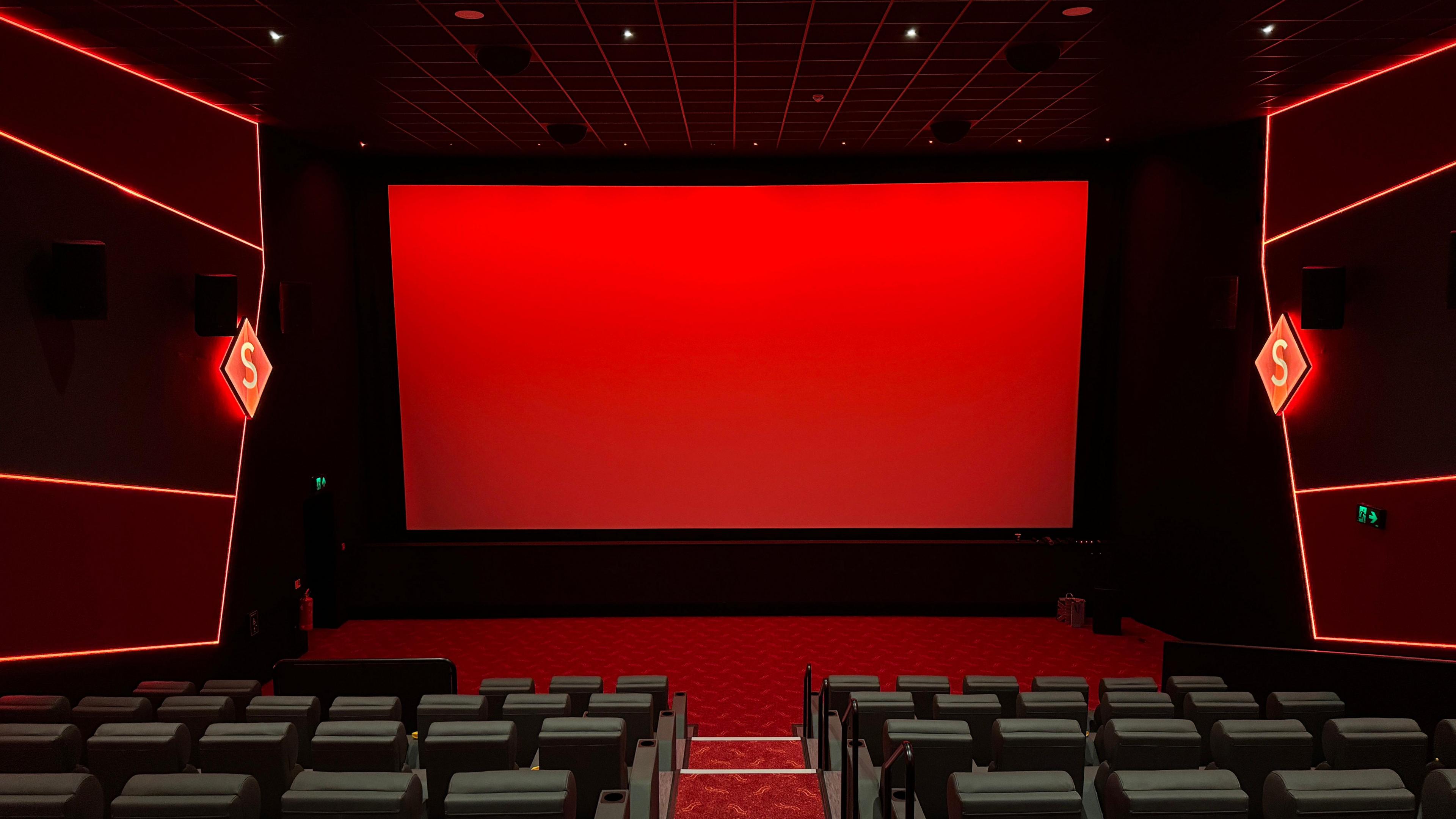 A view from inside a cinema room. There are rows of black leather seats and a red screen at the front. There is red branding down the sides