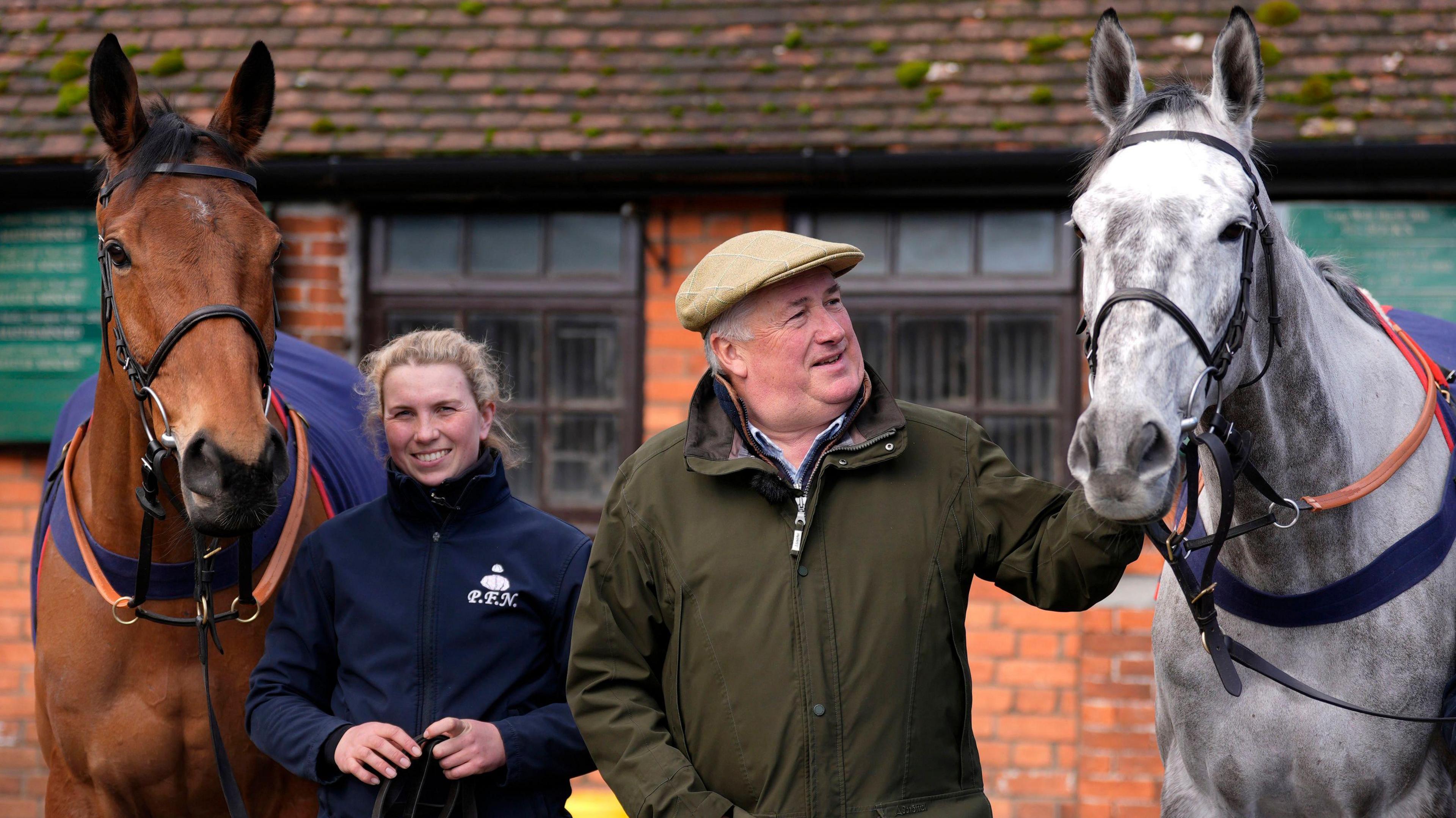 Two people are standing next to each other, framed by two horses. The left horse is brown, the horse to the right is white. In the background is a farm house.