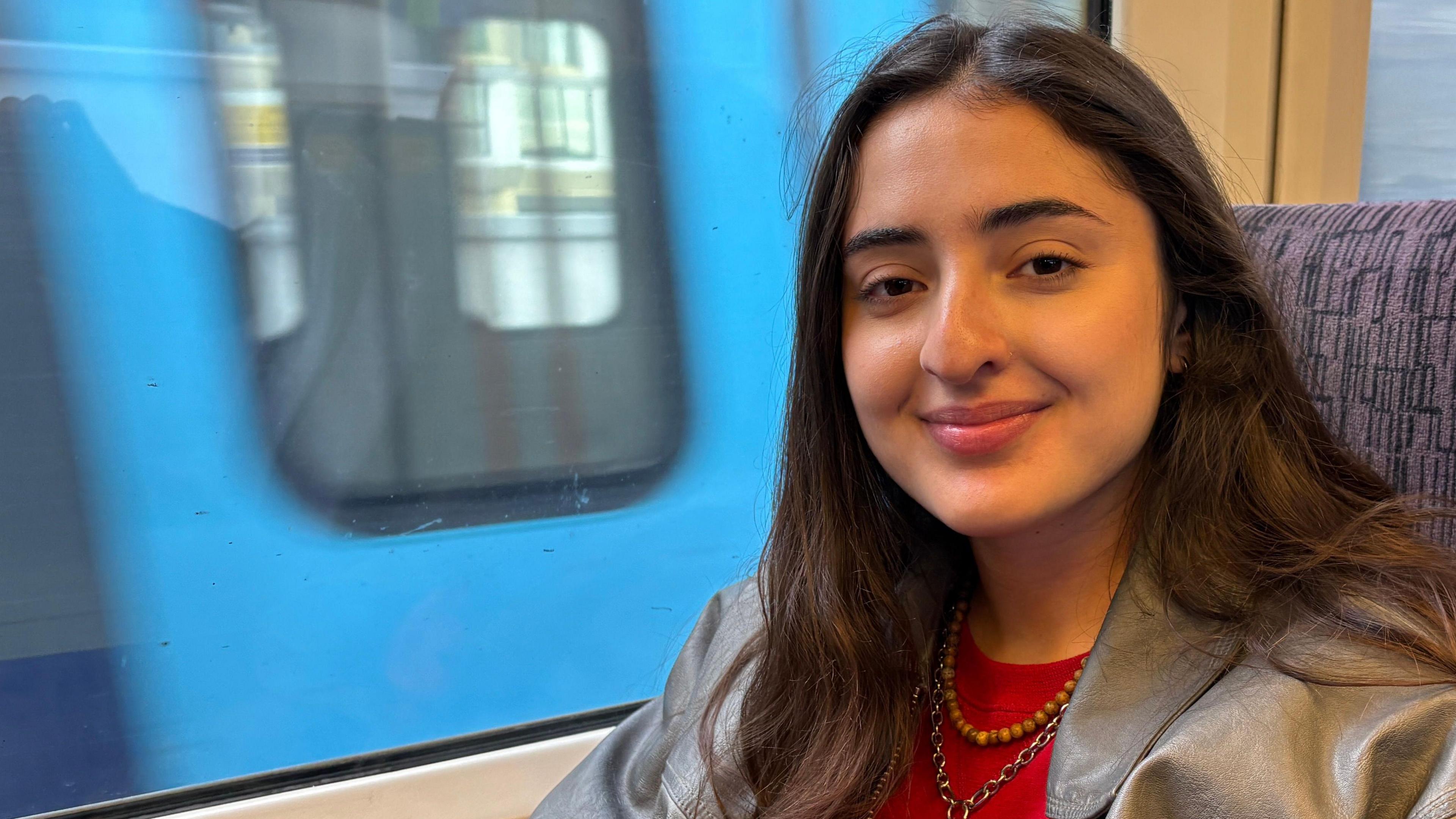 Tala Yunis looking directly down the camera. She is smiling, though her teeth are not visible. She has long brown straight hair that is falling over her shoulders. She is wearing a grey-green coat over a red top, as well as two necklaces. She is sitting on a train, next to a window. The seat back behind her is  purple and patterned. Through the window, a blue train can be seen travelling in the other direction; it is blurred, giving the impression of movement. 
