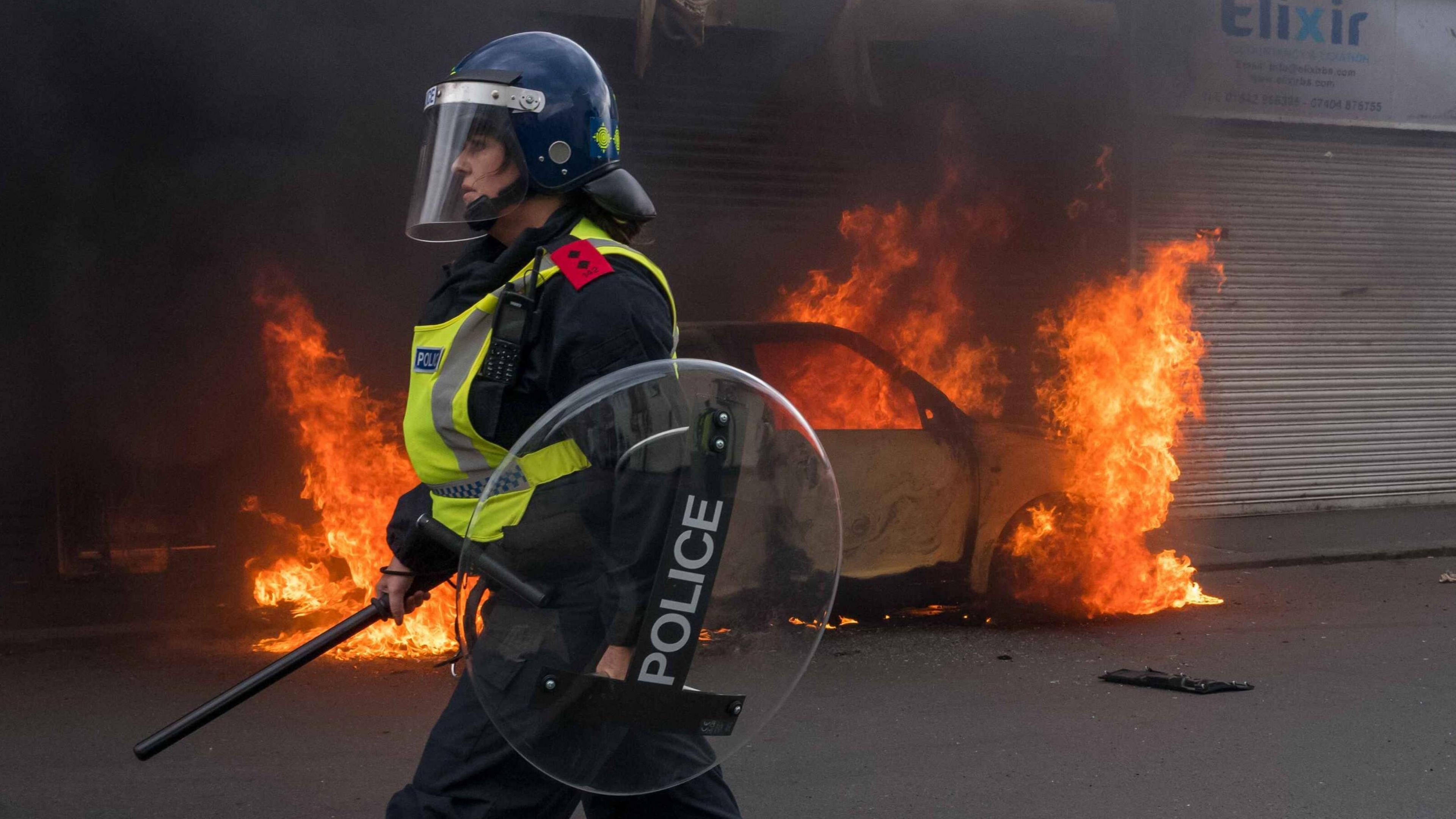 A car burns on a road during a riot. A police women , wearing riot clothing and a shield is standing in front of it.