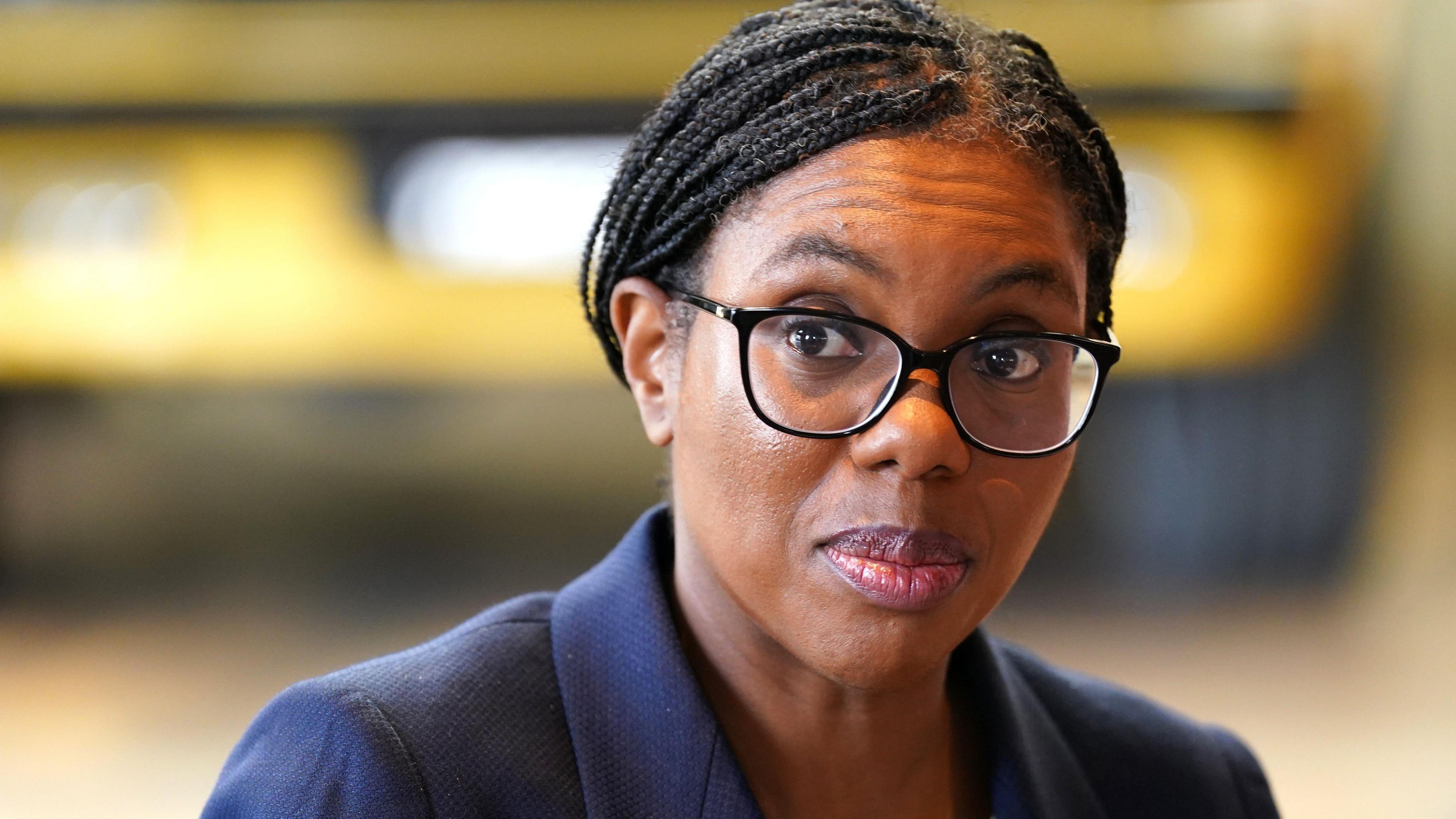A picture of Kemi Badenoch from the shoulders up. She is wearing a dark blue suit jacket and black-rimmed glasses. Her hair, which is in braids, is tied back. She is looking to the left and the background is blurred.