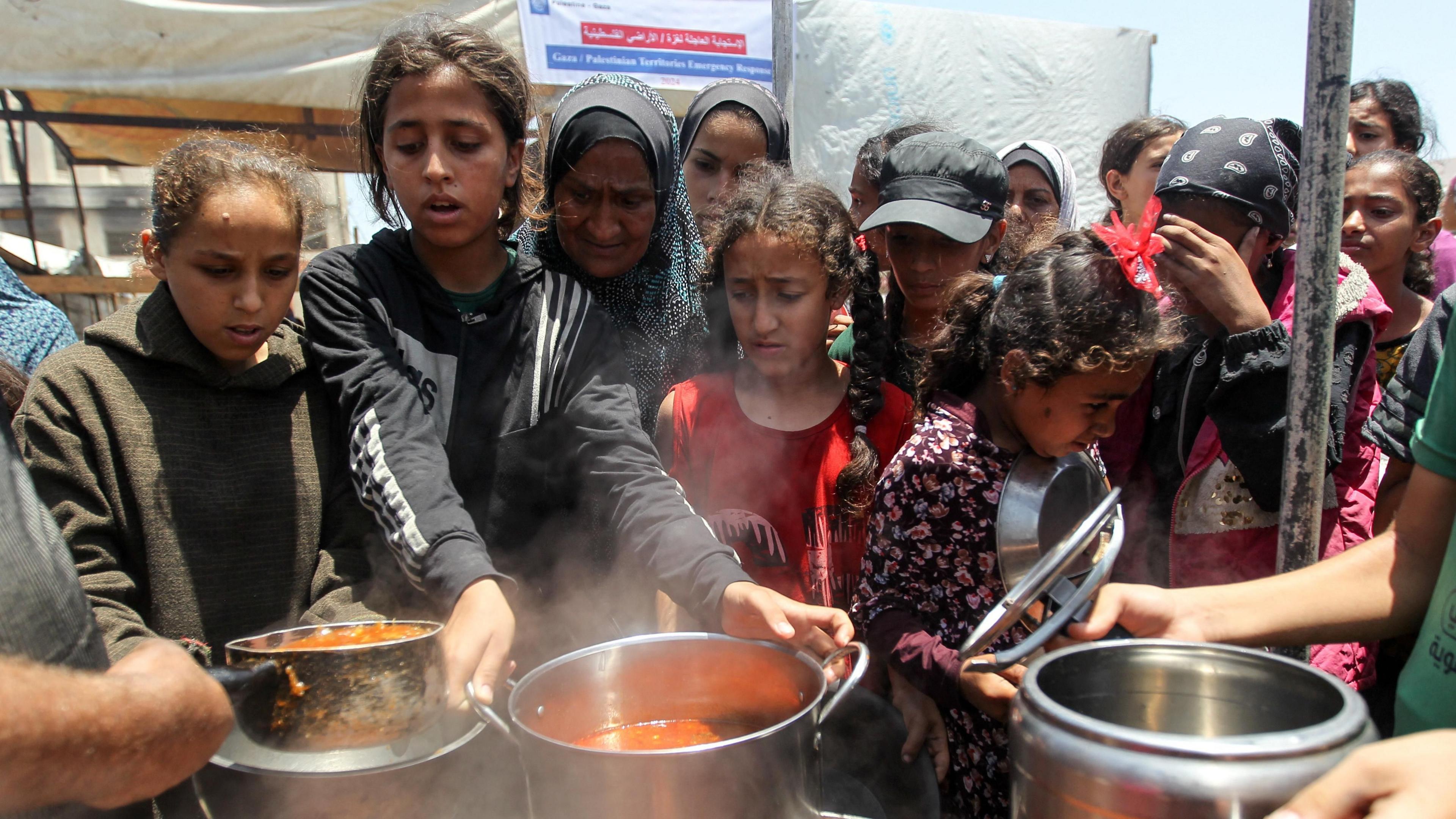 Young Palestinian women and girls queue for aid, pots of food are cooking at what looks to be a soup station - there is a group of 10 people visible, all female, some wearing head coverings, and all with serious expressions 