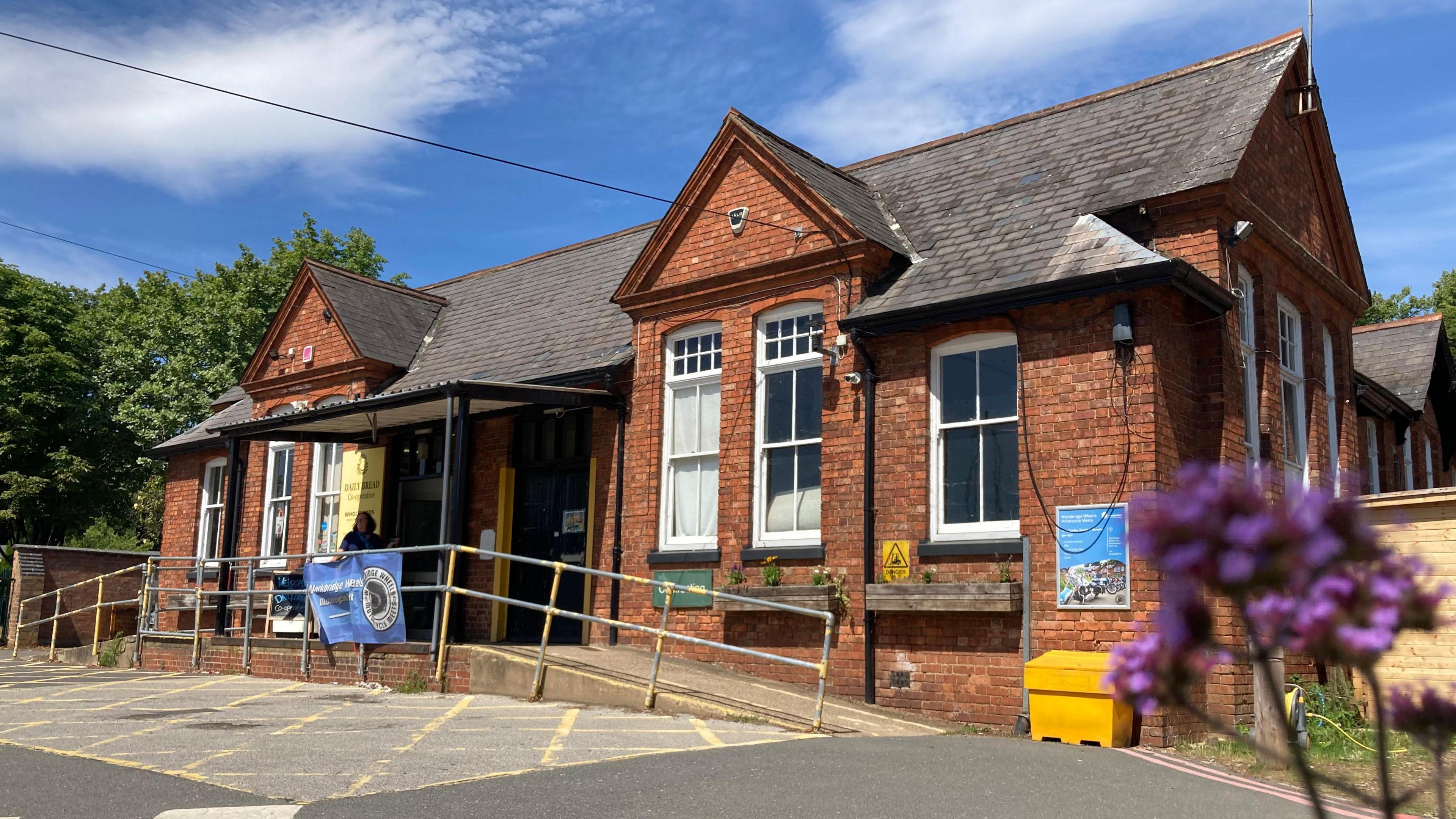 The front of the Workbridge building on Bedford Road in Northampton. It shows a red brick victorian building, much like an old schoolhouse. There are metal railings to the front of the property for wheelchairs to go up a ramp. In the foreground, to the right of the picture, is a purple verbena which is blurred. There is a yellow grit bin outside the front of the building, which has large white sash windows.