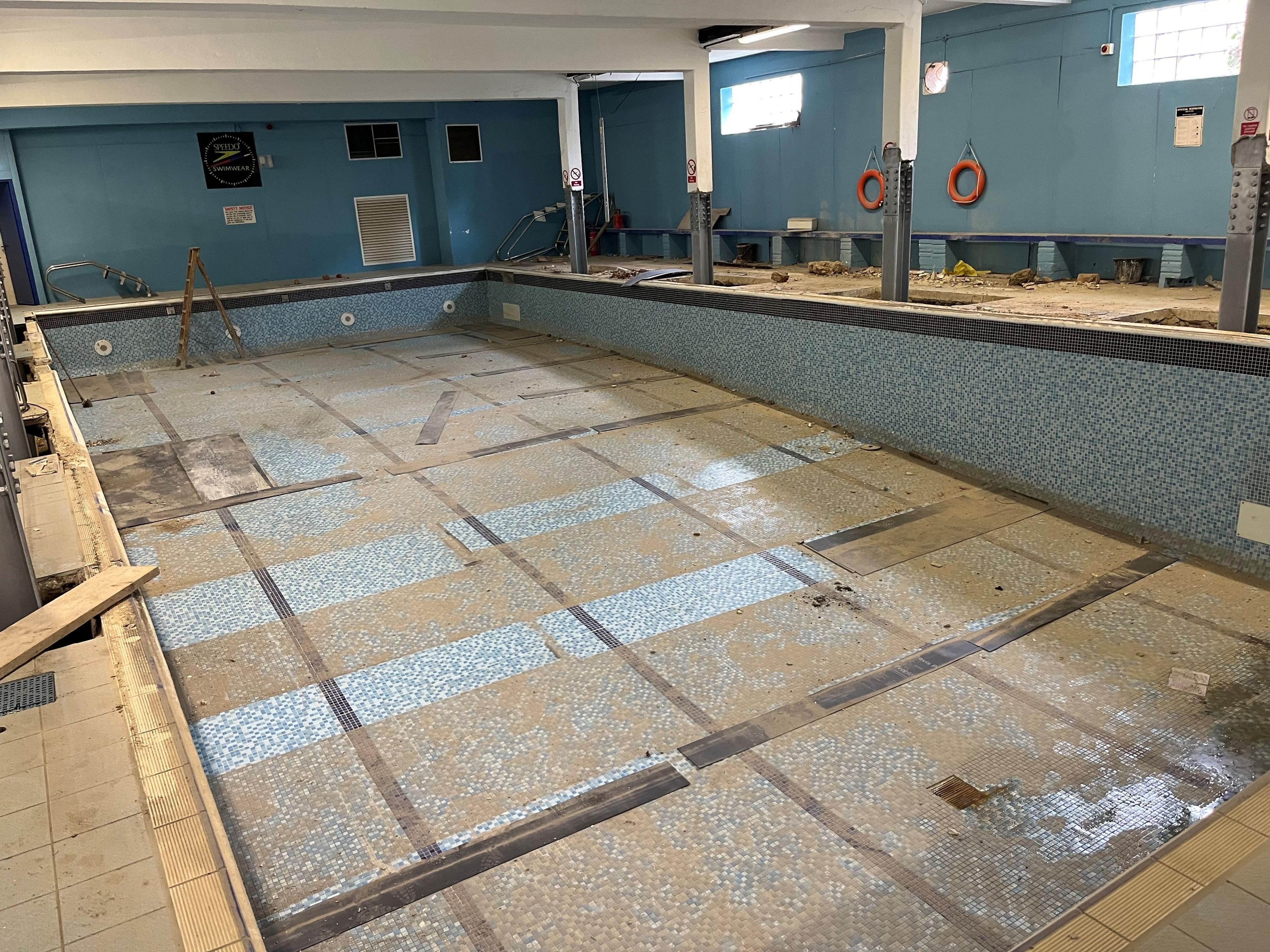 An empty swimming pool, roughly 17m long (55ft) and 8m (26ft) wide. It has blue and white tiles on the sides and bottom of the pool area, with no water in it but lots of brick dust with building materials seen around the outside.