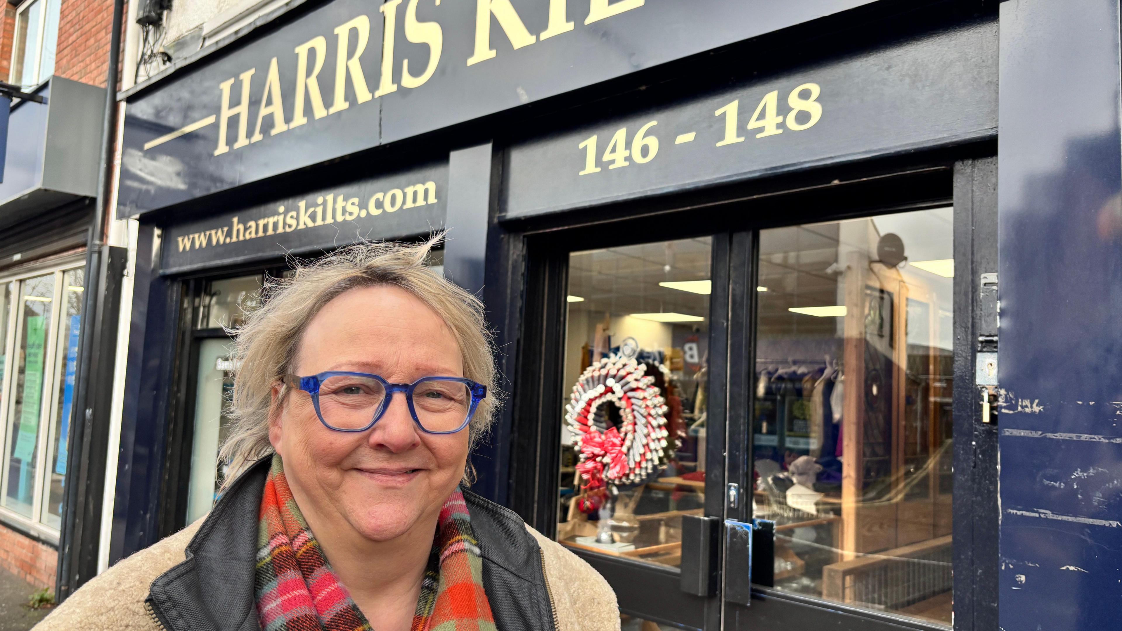 Kim Harris. She has blonde hair, wearing glasses, a beige coat with black collar. A blue shop-front for Harris Kilts is behind her.