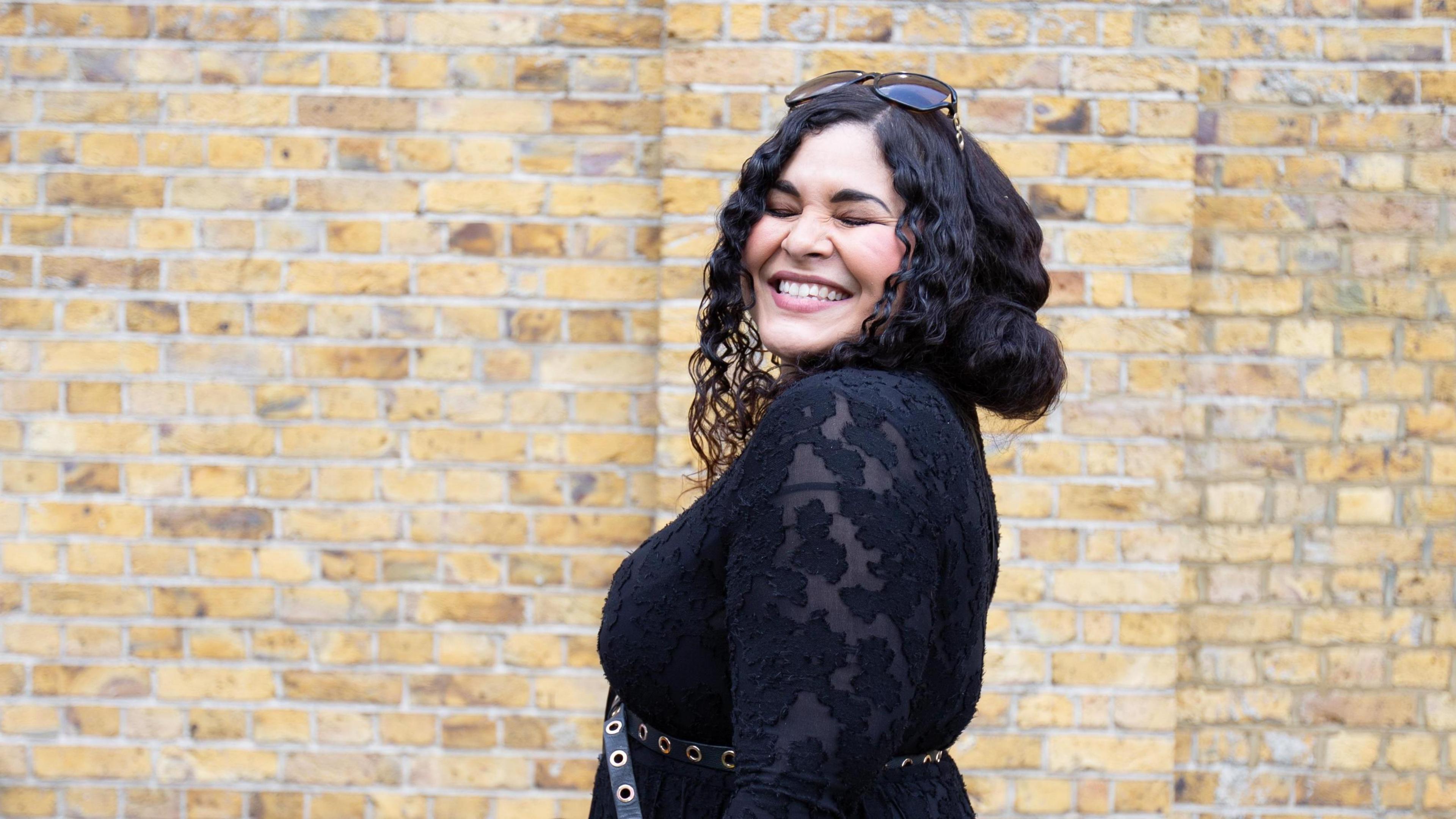 Gemma Devonish, a woman with dark curly hair, wearing sunglasses on her head and smiling broadly with her eyes shut