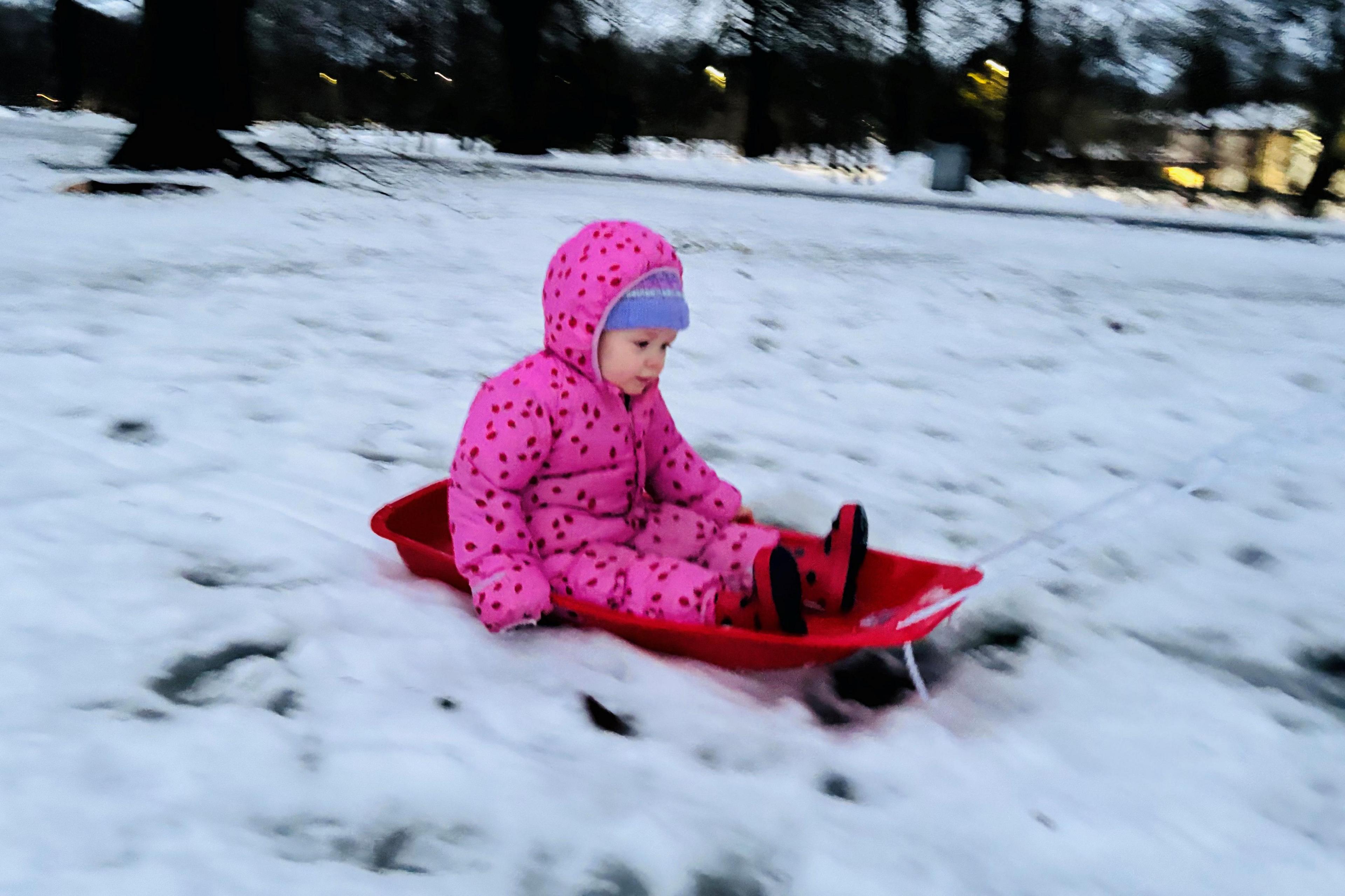 Two-year-old girl in bright pink snowsuit on a red sledge in a snow-covered park.