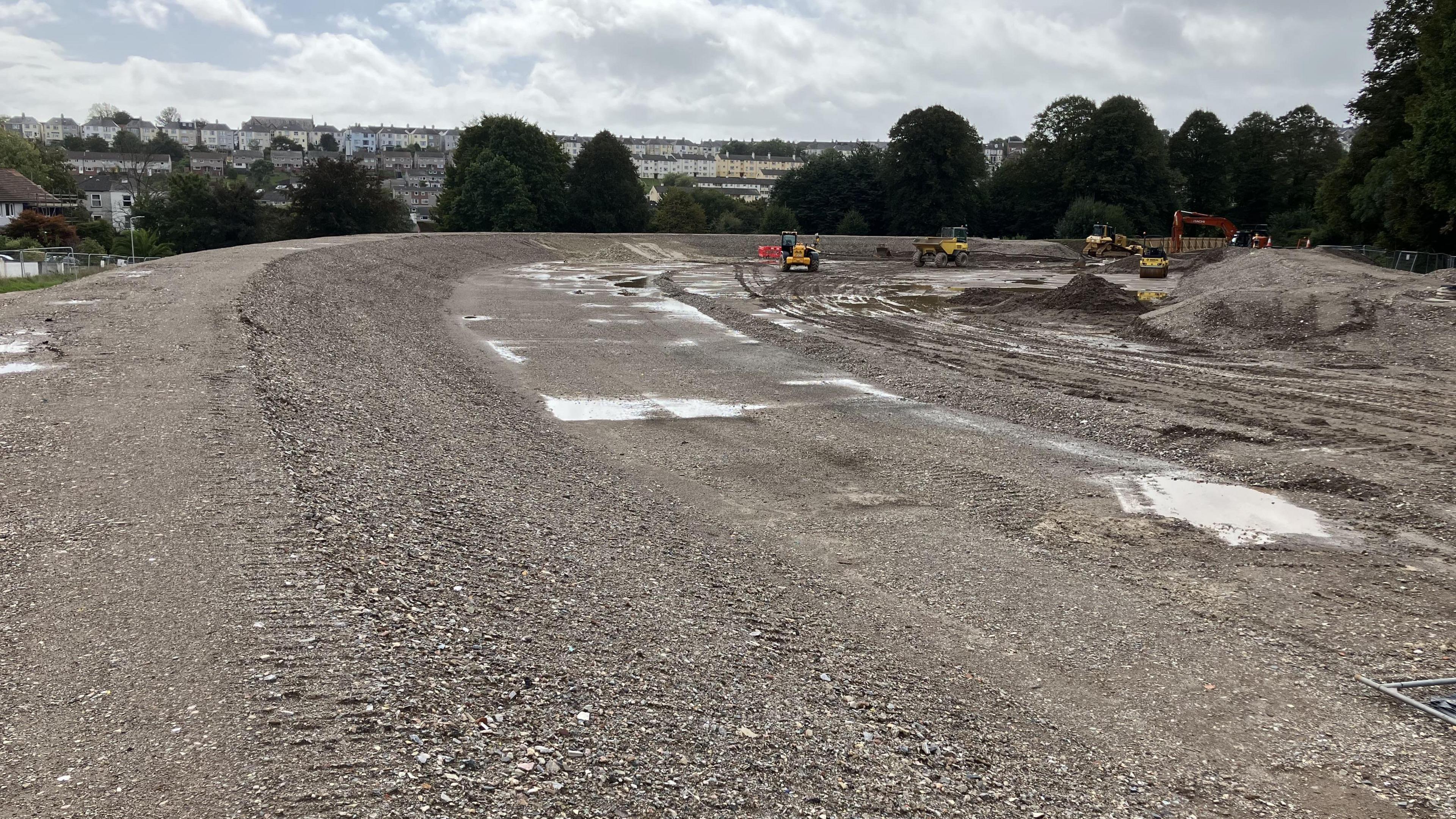 A picture of large embankments that are being built on an open area with the city of Plymouth.  There are yellow diggers moving earth in the background - to create banks that will hold back flood water.  The sky is overcast and there are puddles of water on the rocky soil.  