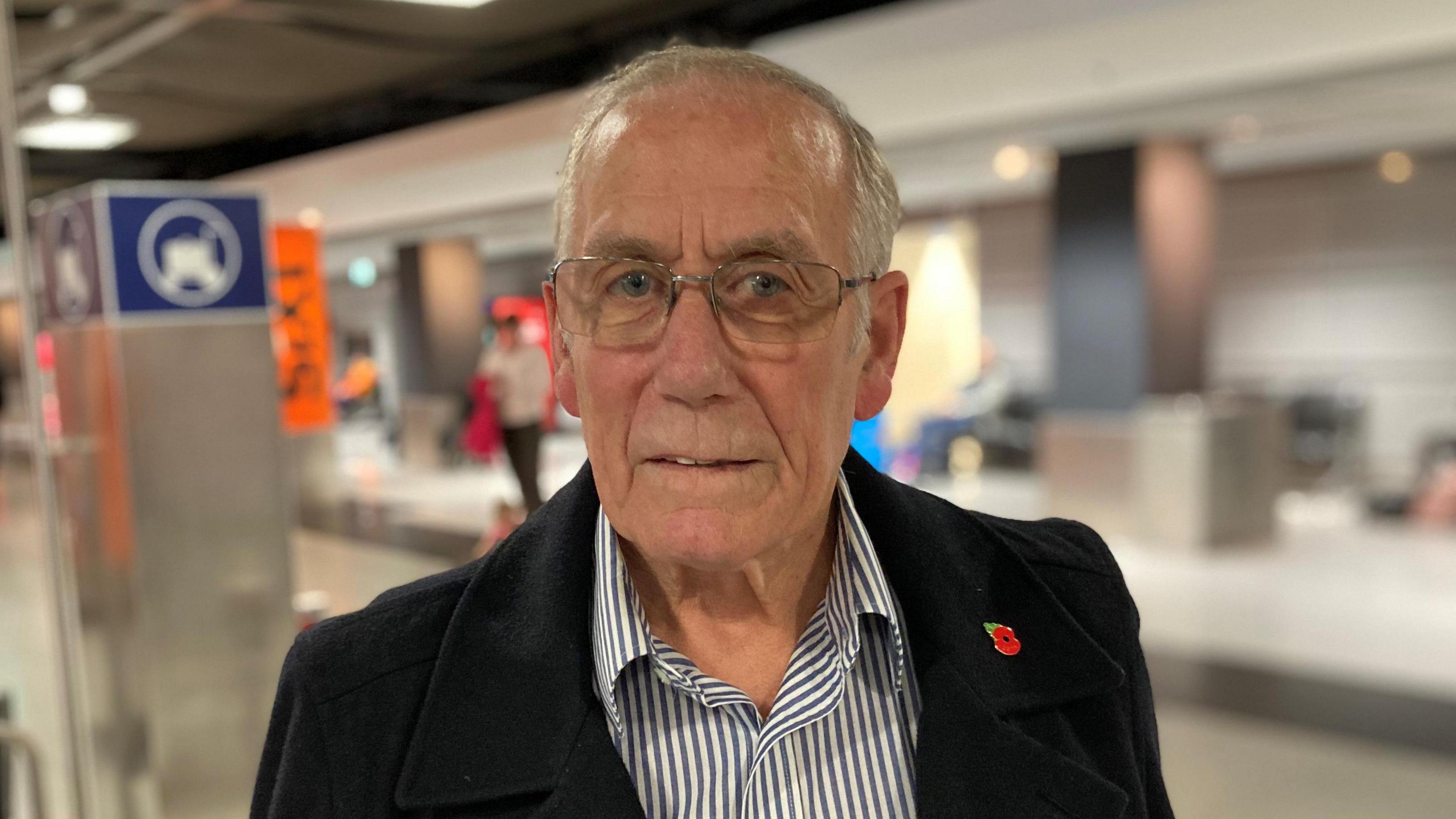 Richard Lee at an airport in Germany. He is wearing a white and blue striped shirt and a black coat with a metal poppy pin in his lapel. He has short grey hair and glasses.