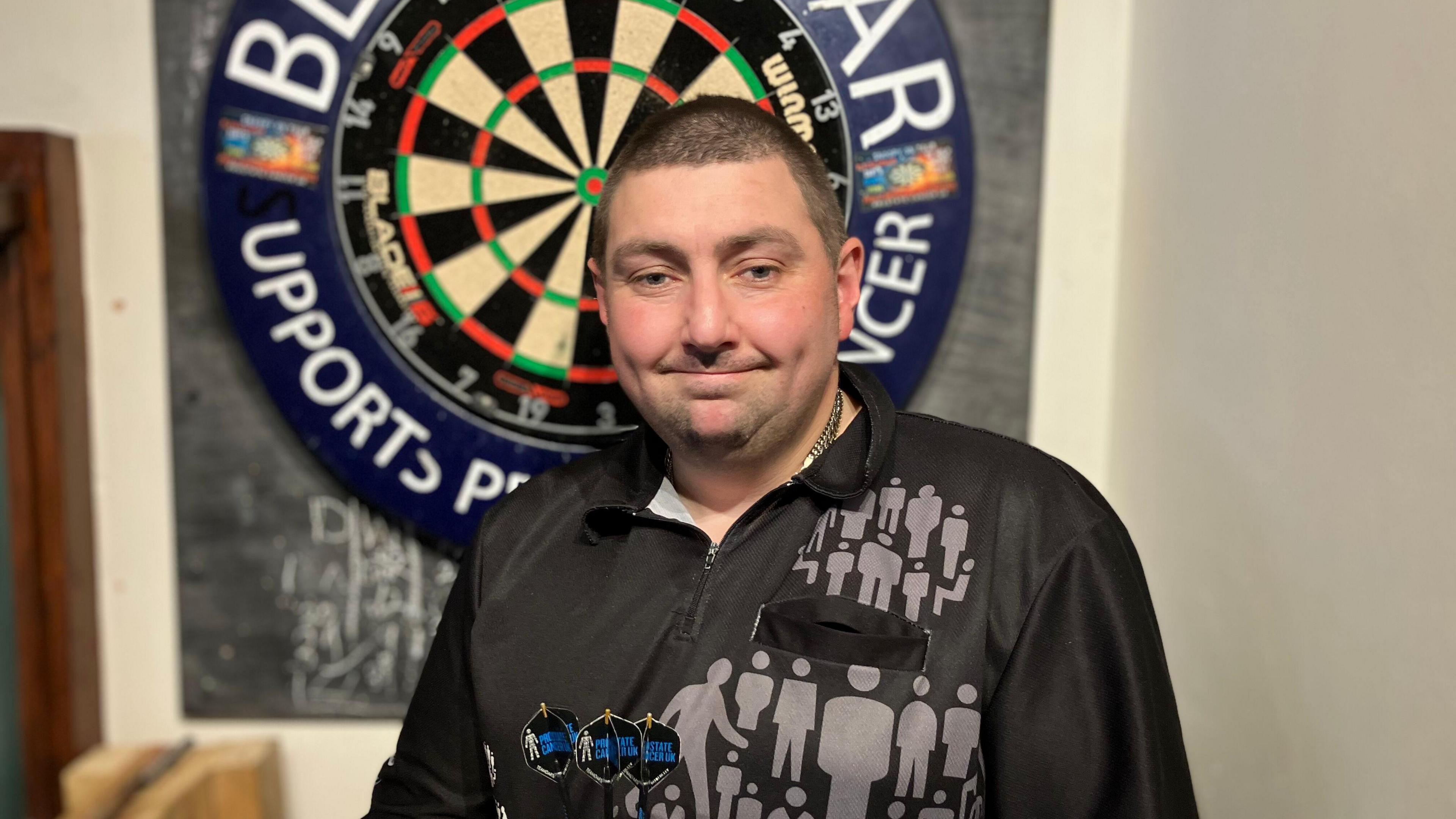 A man with black hair in a short buzz cut style. He is in a black darts shirt and holding three darts that have the Prostate Cancer UK logo on the flights. He is stood in front of a darts board.