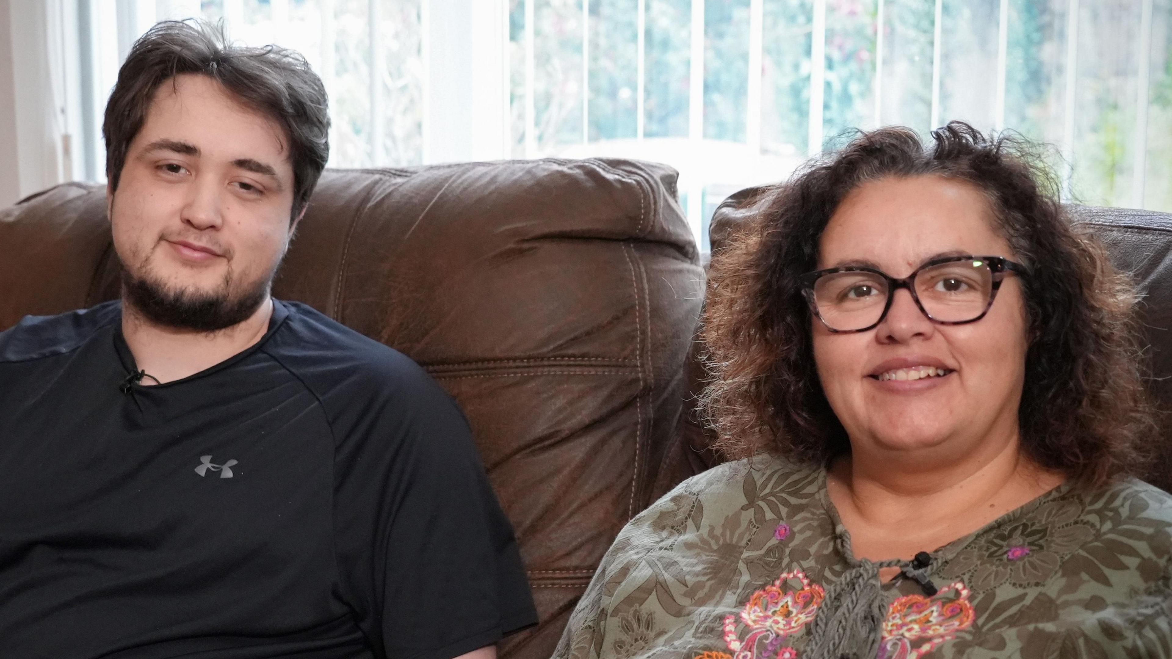Tomas Cozens and his mum Jo sitting next to each other on a brown leather sofa. Tomas is on the left and is wearing a black Under Armour t-shirt and has dark wavy hair and a brown goatee beard. Jo has curly brown hair down to her shoulder and has a green floral print top on that has laces on the chest. It is a head and shoulders shot of them.