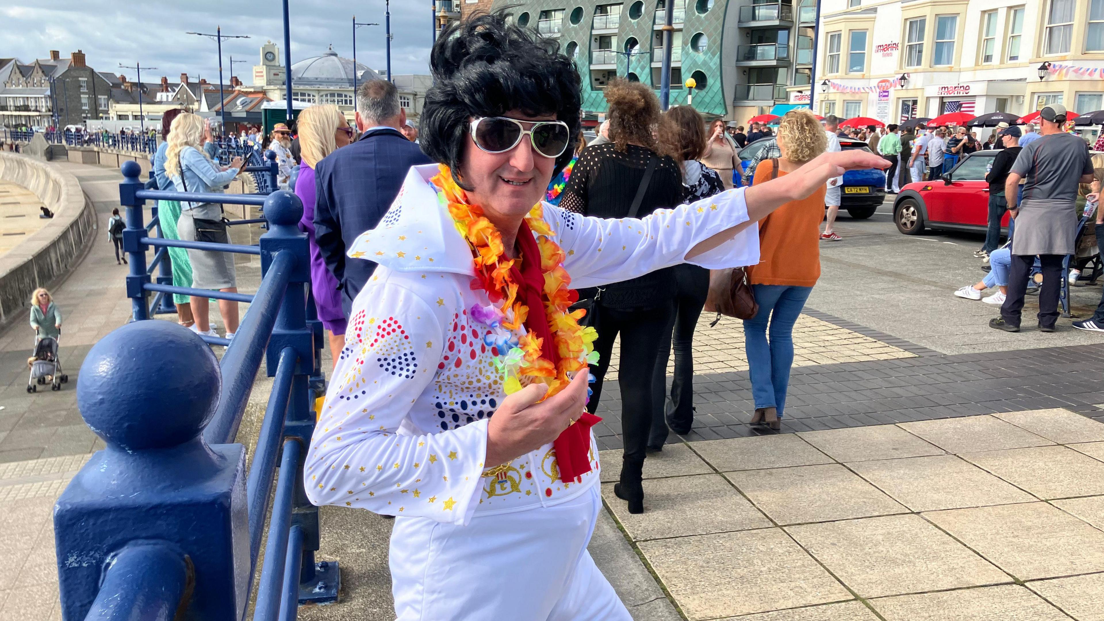 An Elvis in a white outfit smiles and strikes a pose, pointing with his left hand