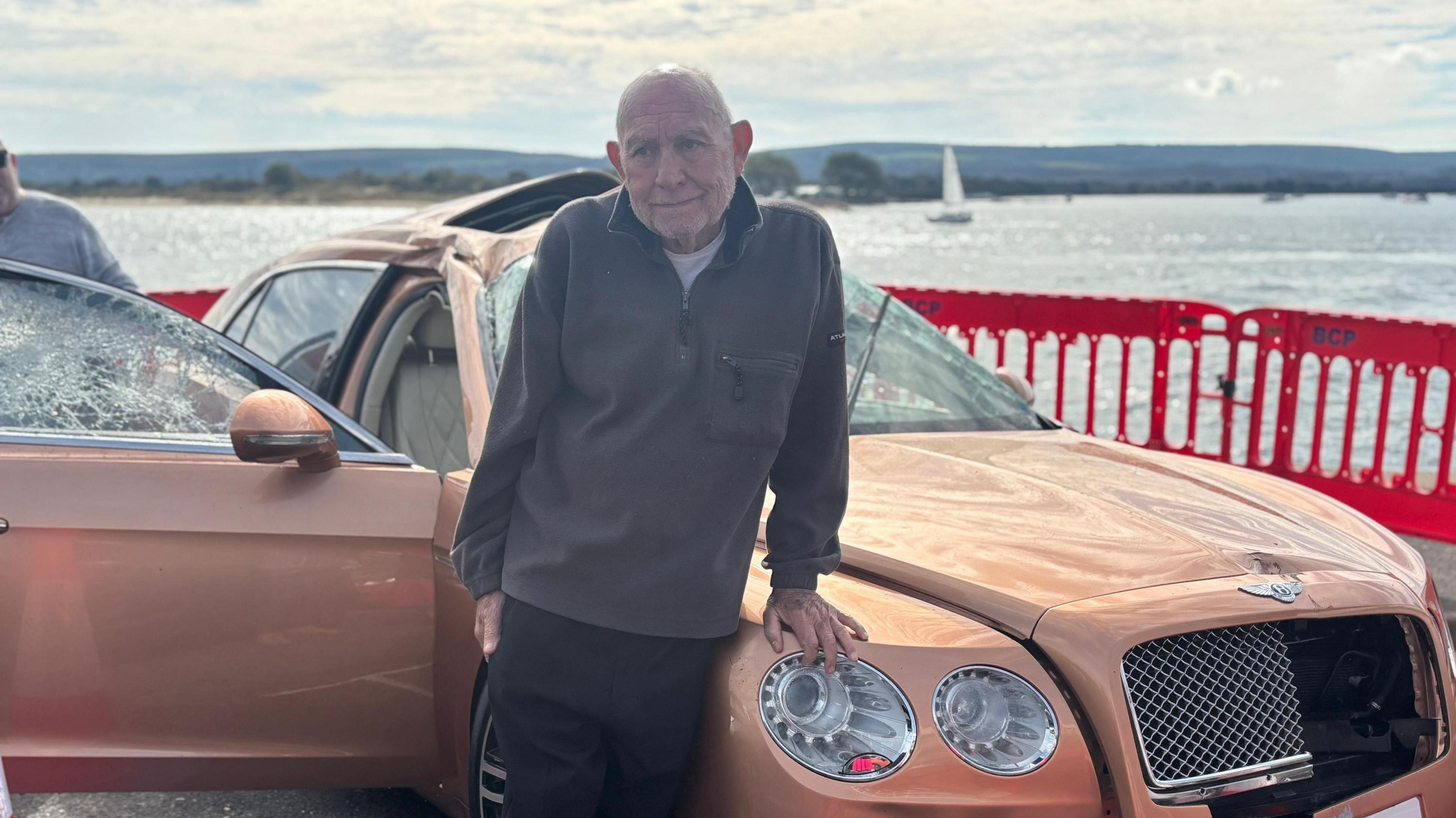 A bronze Bentley car with a crumpled roof and broken windscreen after being lifted from the seabed. A man in his 70s is in front and leaning against it.