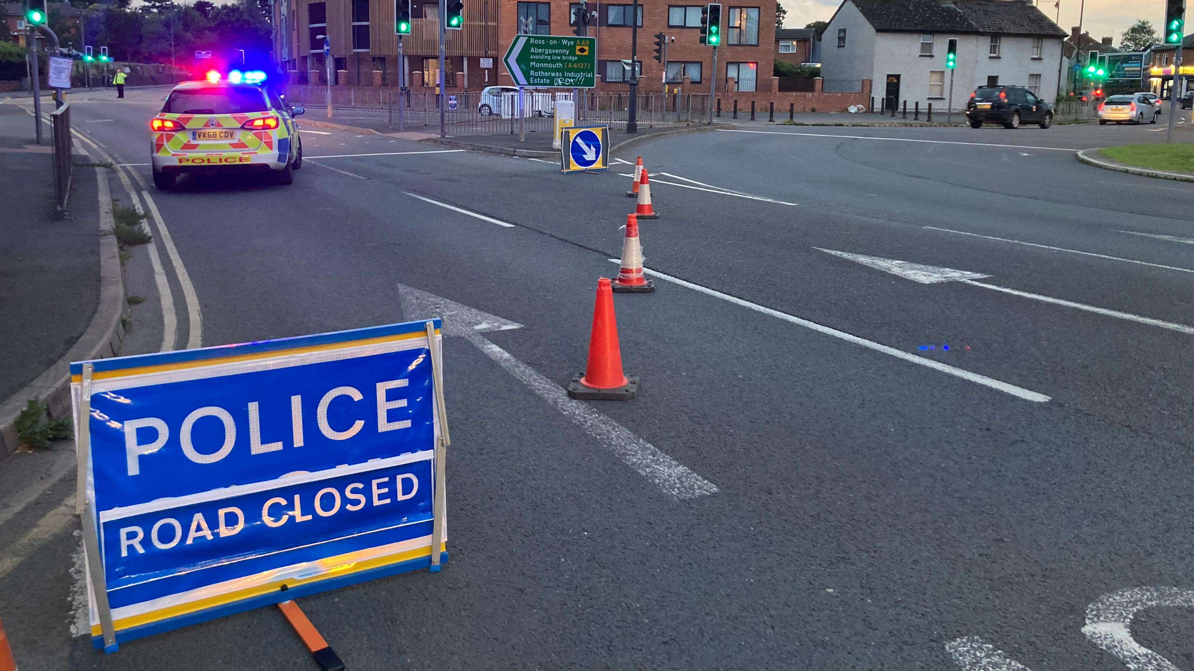 A blue sign with white writing that reads " POLICE ROAD CLOSED" and a police car with it's lights on in the background while a road is closed off by police tape