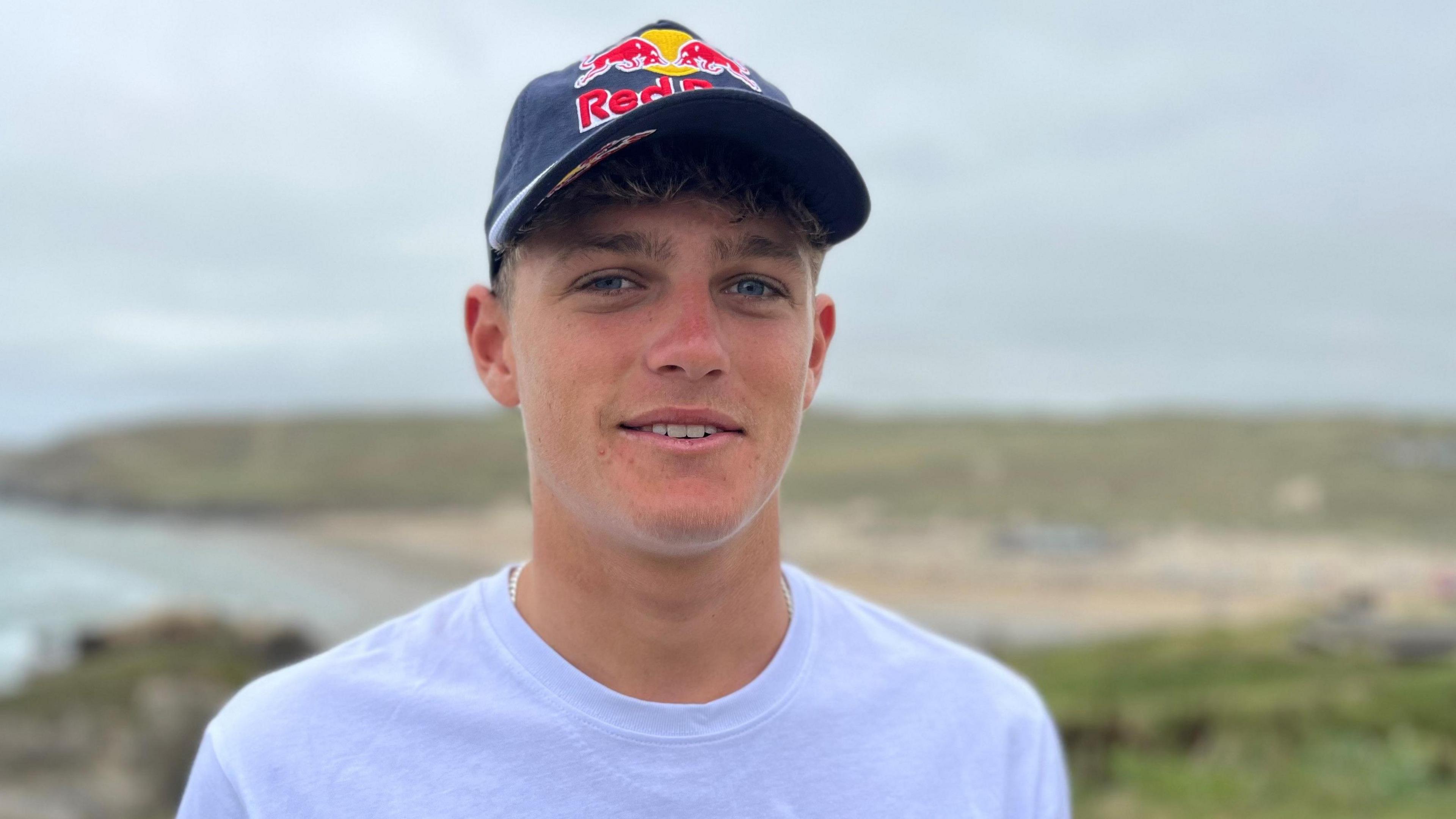 A teenage boy with blue eyes is looking directly at the camera and smiling, he is wearing a blue cap that says Red Bull and a white t-shirt. He is standing on a cliff overlooking a beach that is blurred in the background