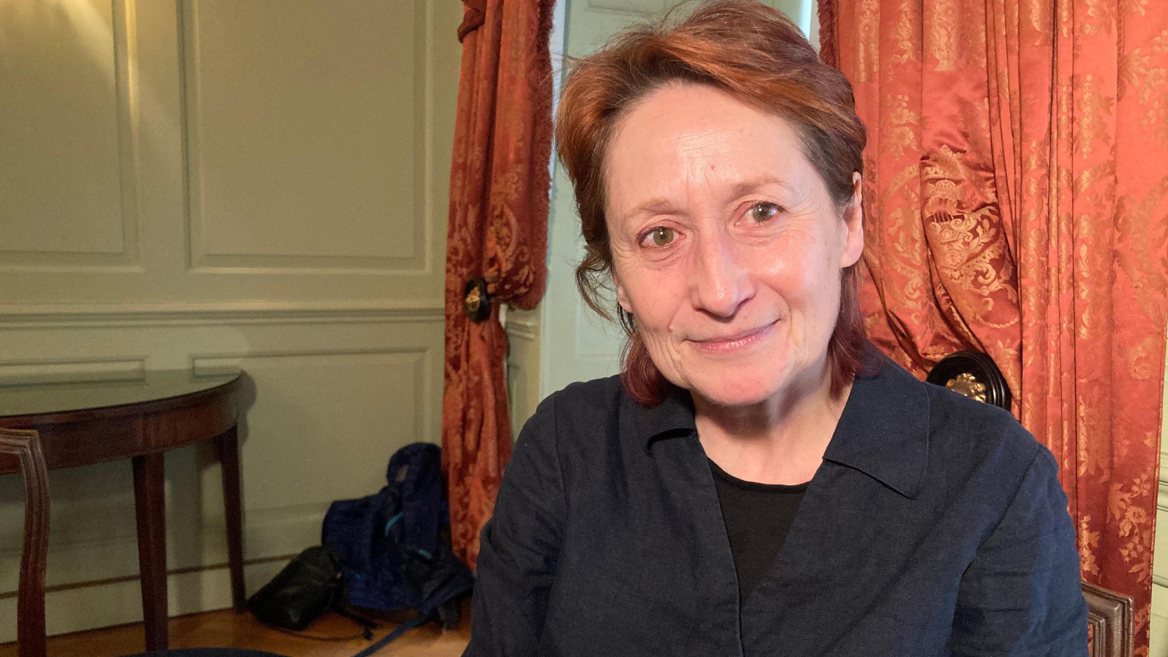 Prof Jennifer Richards is sitting inside a room with some large grand curtains hanging behind her. She has shoulder length red hair, which has been tucked behind her ears. She has green eyes and is wearing a blue shirt. She is also smiling at the camera. 