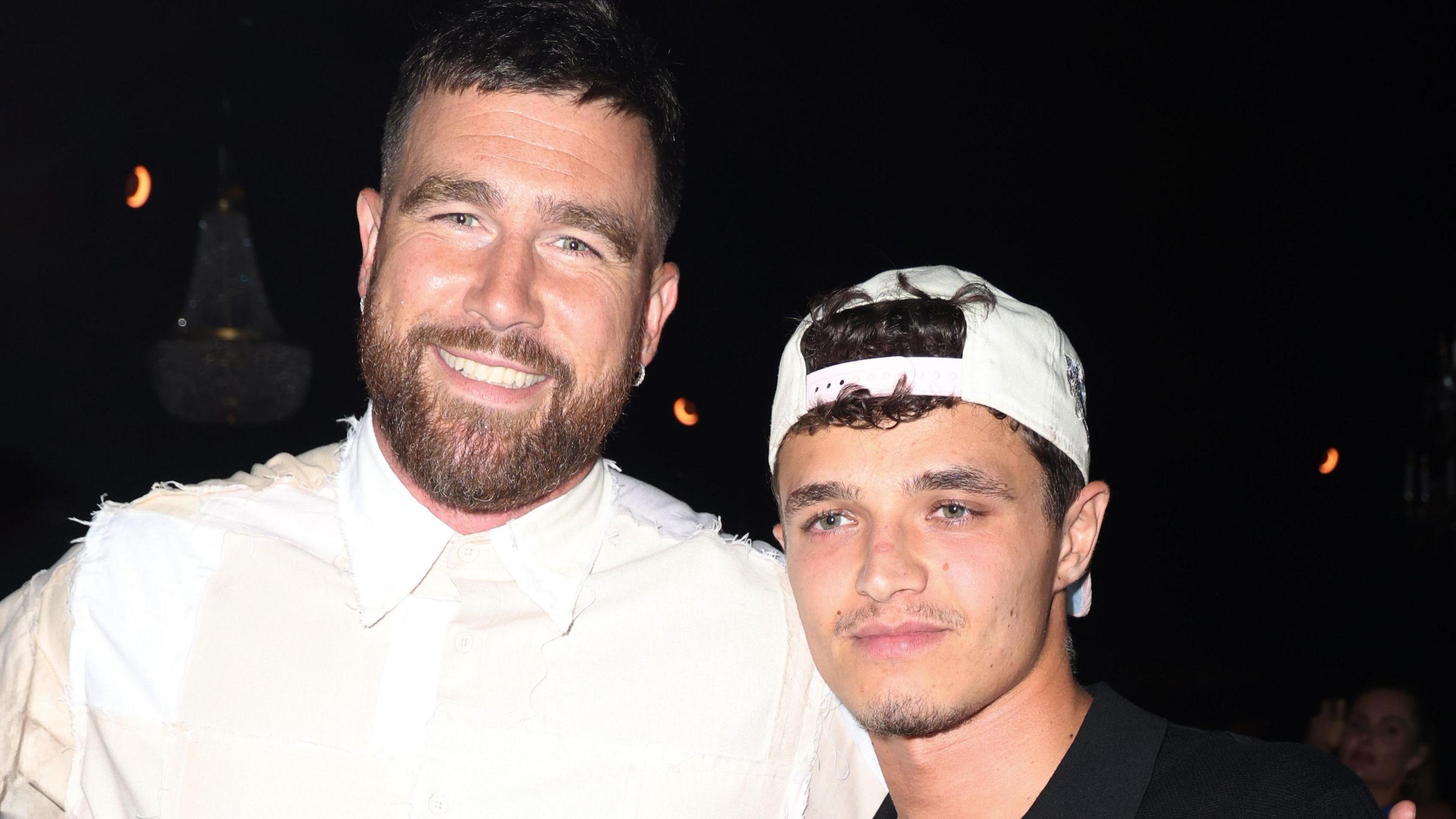 Travis Kelce and Lando Norris at a party in Miami on 5 May. Travis is a white man in his 30s with blue eyes, short brown hair and a short brown beard. He smiles at the camera, wearing a white shirt. Lando is a 24-year-old white man with green eyes and a short moustache and goatee beard. He wears a white cap twisted back over his curly brown hair. He stands about a foot shorter than Travis, who has his arm around him. 