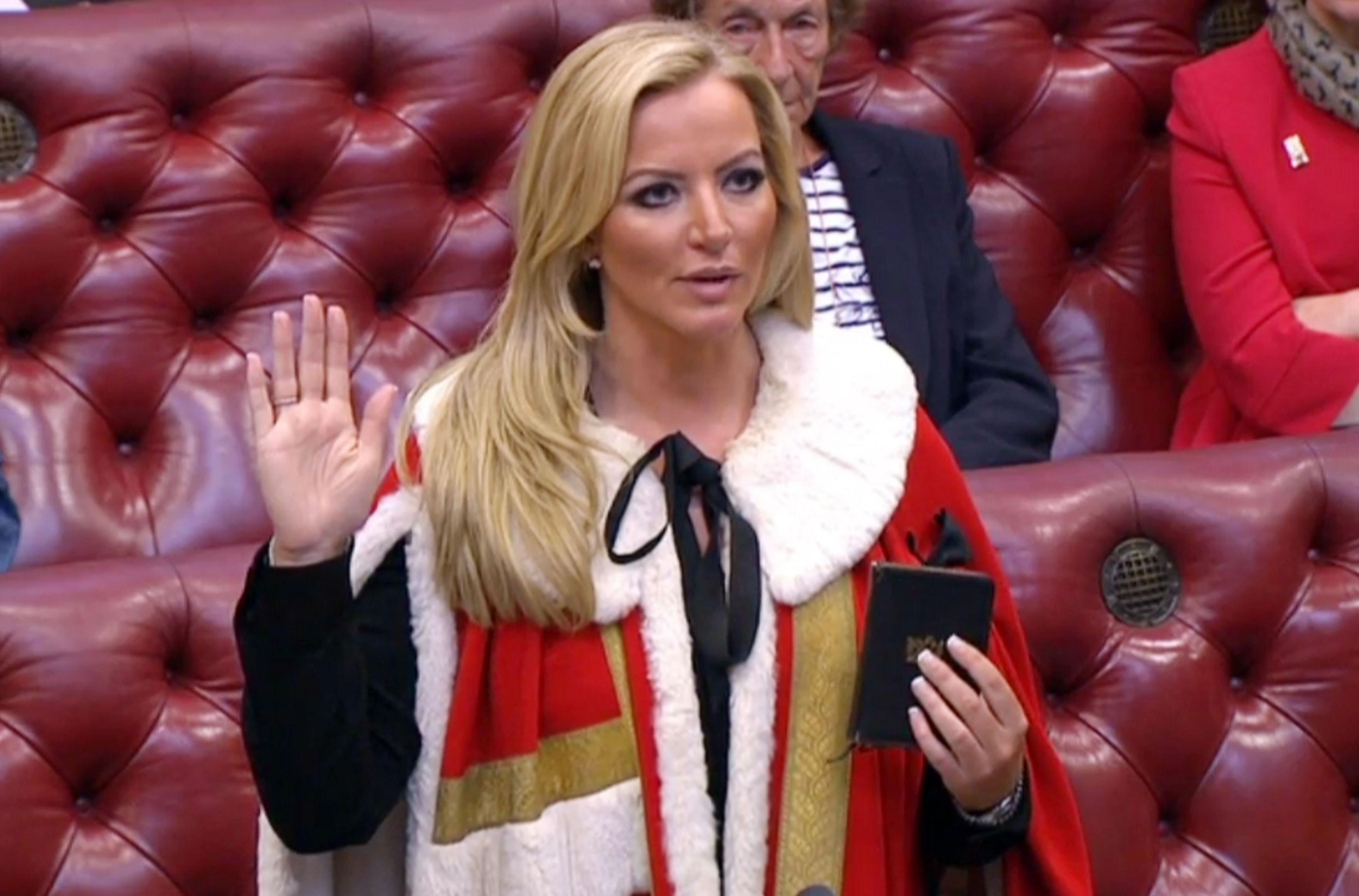 Michelle Mone in red, gold and white ermine and black jacket with a bible in her left hand, holding up her right hand while taking an oath. People can be seen on the red benches behind her.