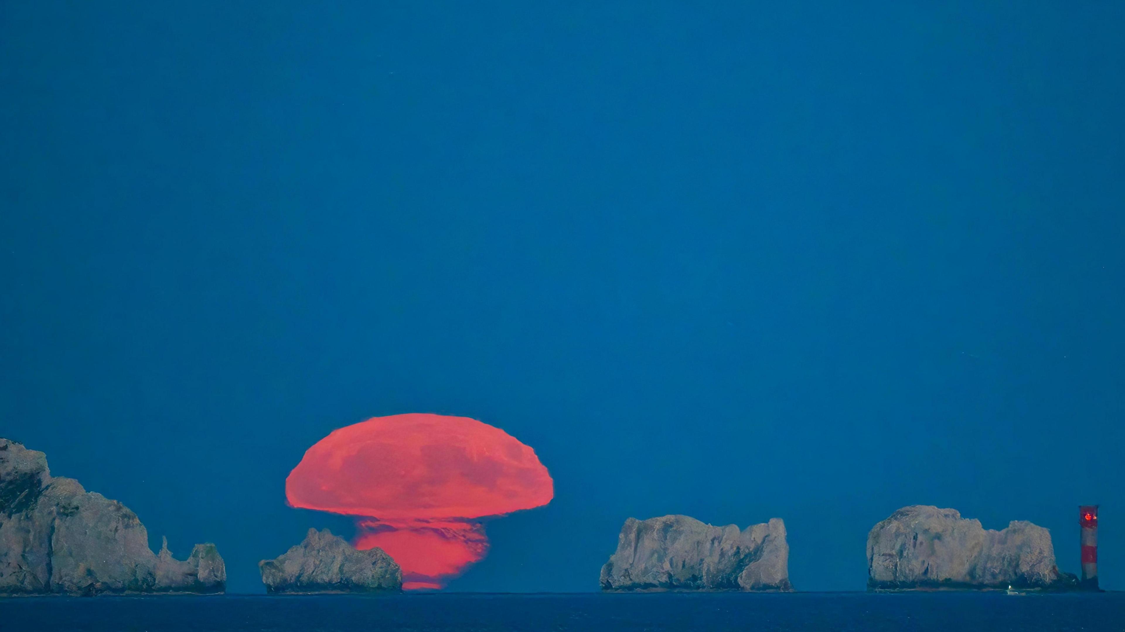 The moon above a rock and cliff formation off the Isle of Wight, a red and white lighthouse to the right. The moon is melding into the horizon like an orange mushroom.